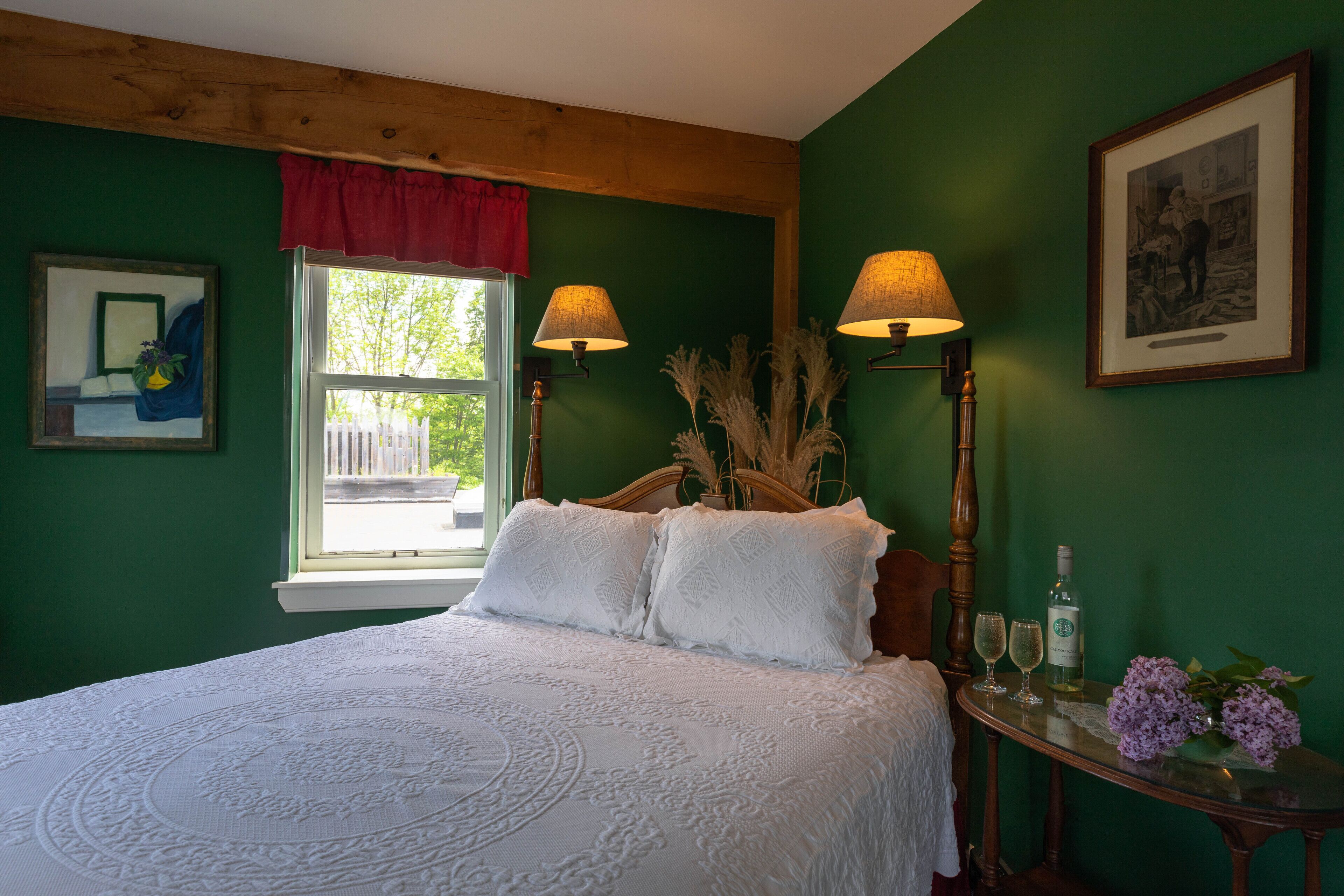 A bedroom with a bed, two wall-mounted lamps, a window with a red valance, a framed picture, and a table with a bottle and flowers.