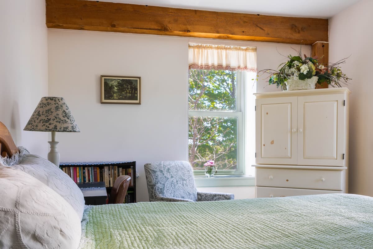 Quarto com cama, cadeira, estante, armário e janela com vista para árvores.