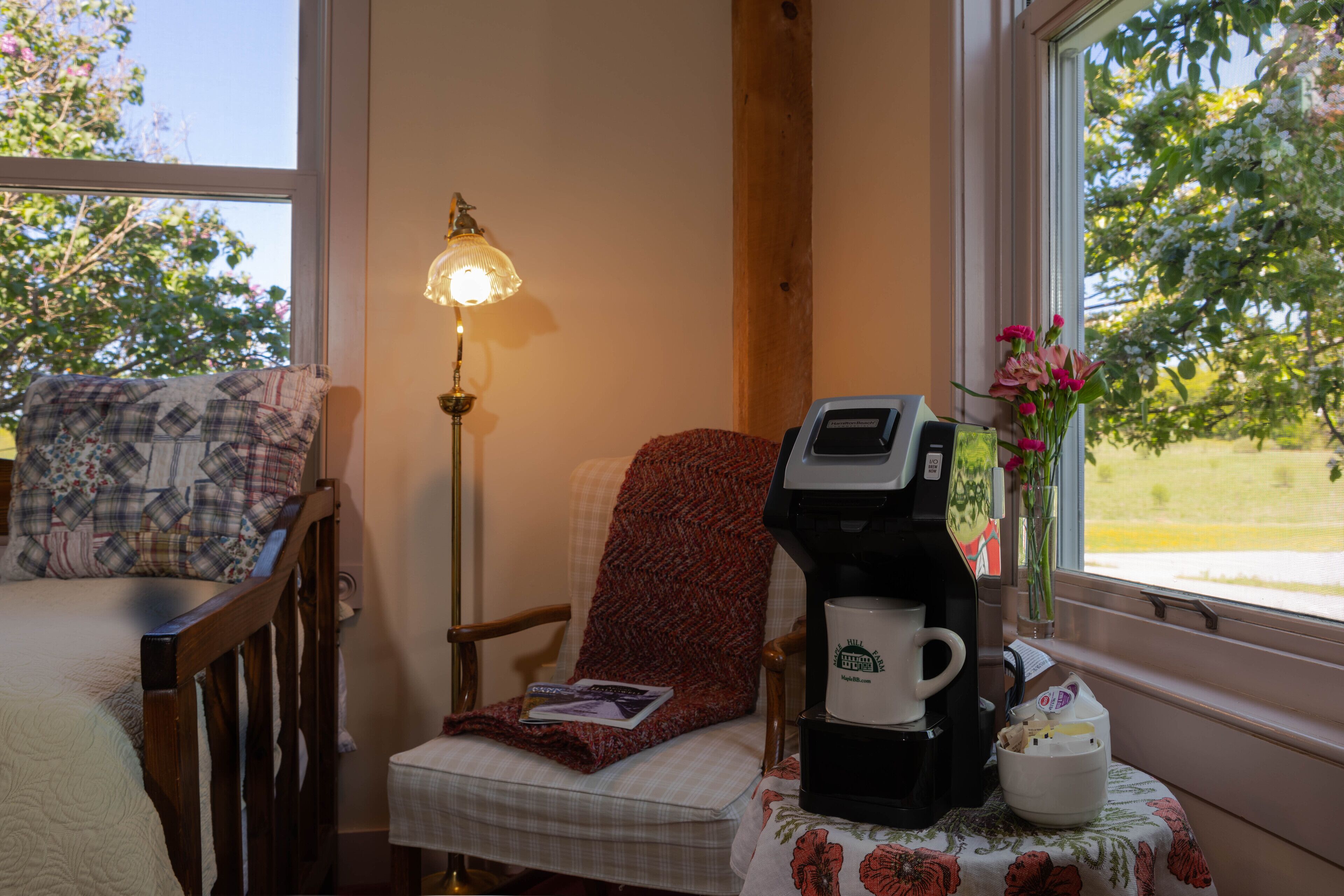 Una habitación acogedora con cama, sillón con manta, cafetera y ventana con vista a árboles.