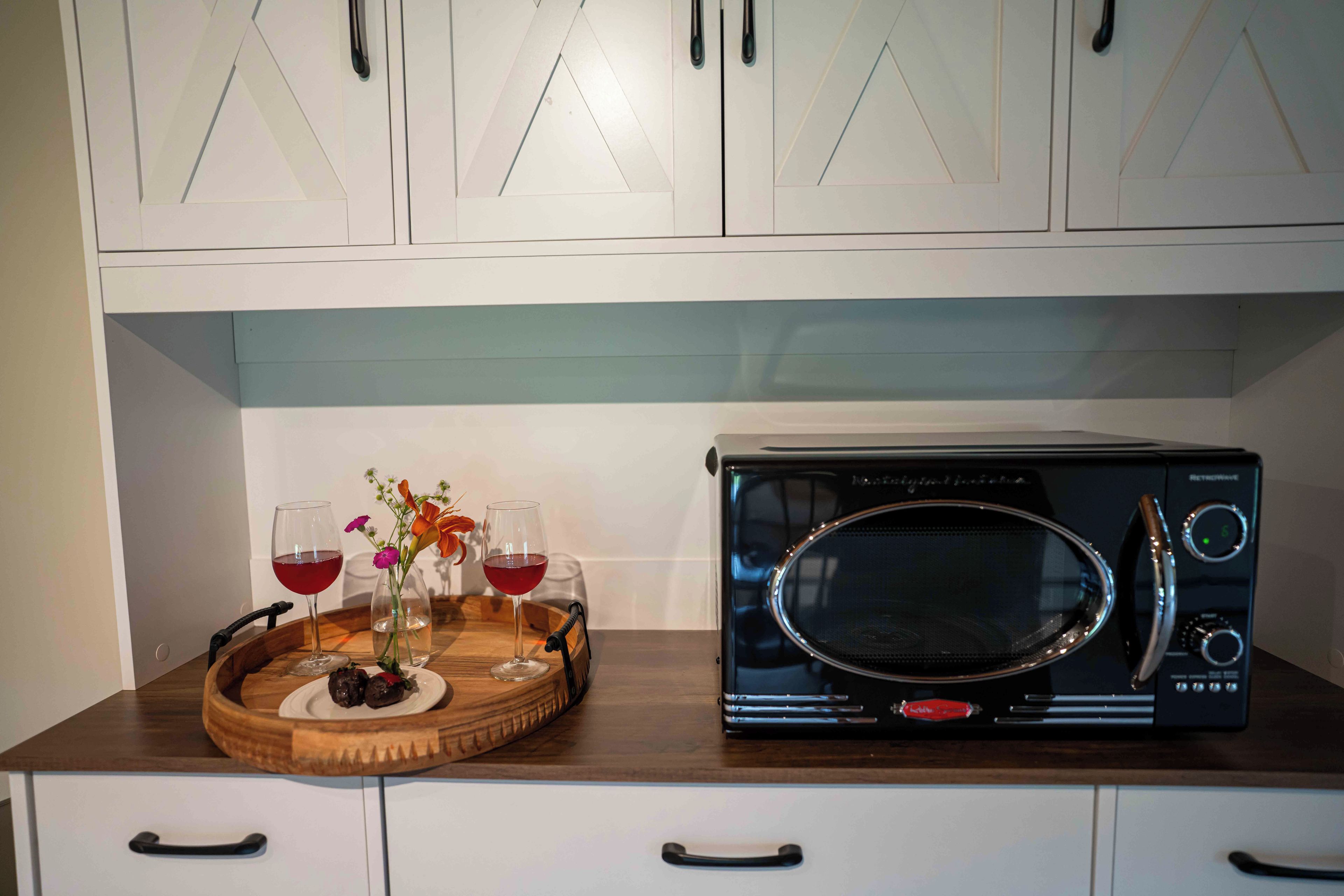 A kitchen with white cabinets, a microwave, a wooden tray with two glasses of red wine and a vase with flowers.