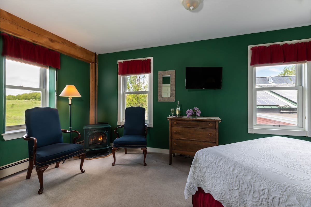 Quarto com paredes verdes, duas poltronas azuis, penteadeira de madeira, cama branca, lareira e televisão na parede.