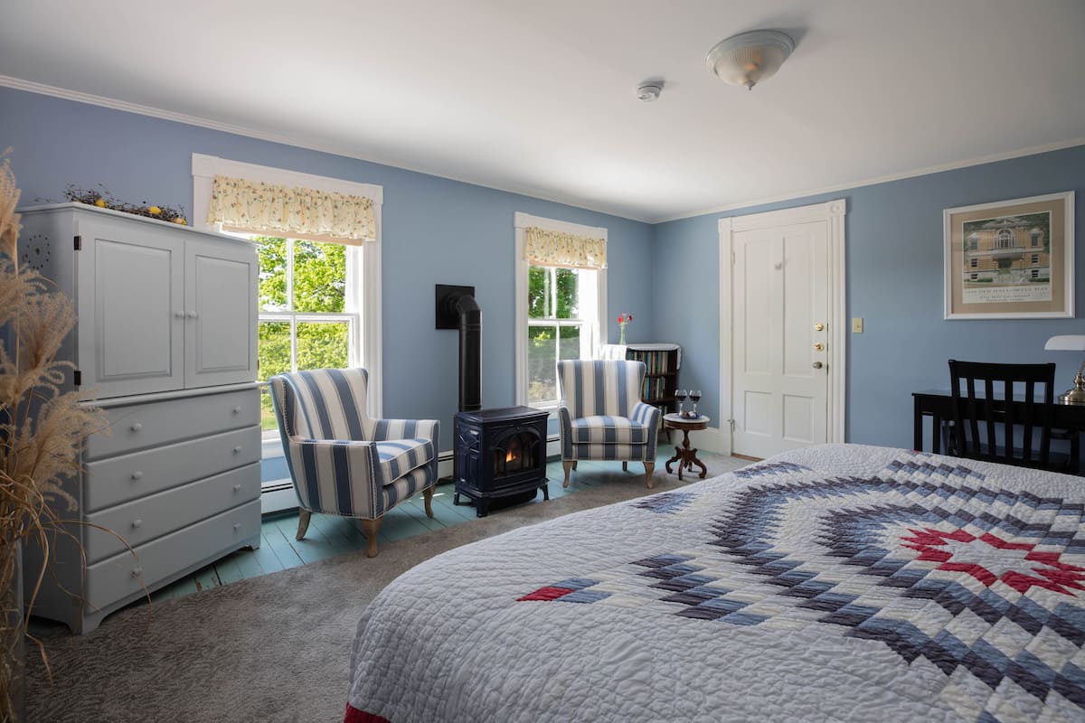 Quarto com cama grande, lareira, duas poltronas listradas, cômoda e estante.