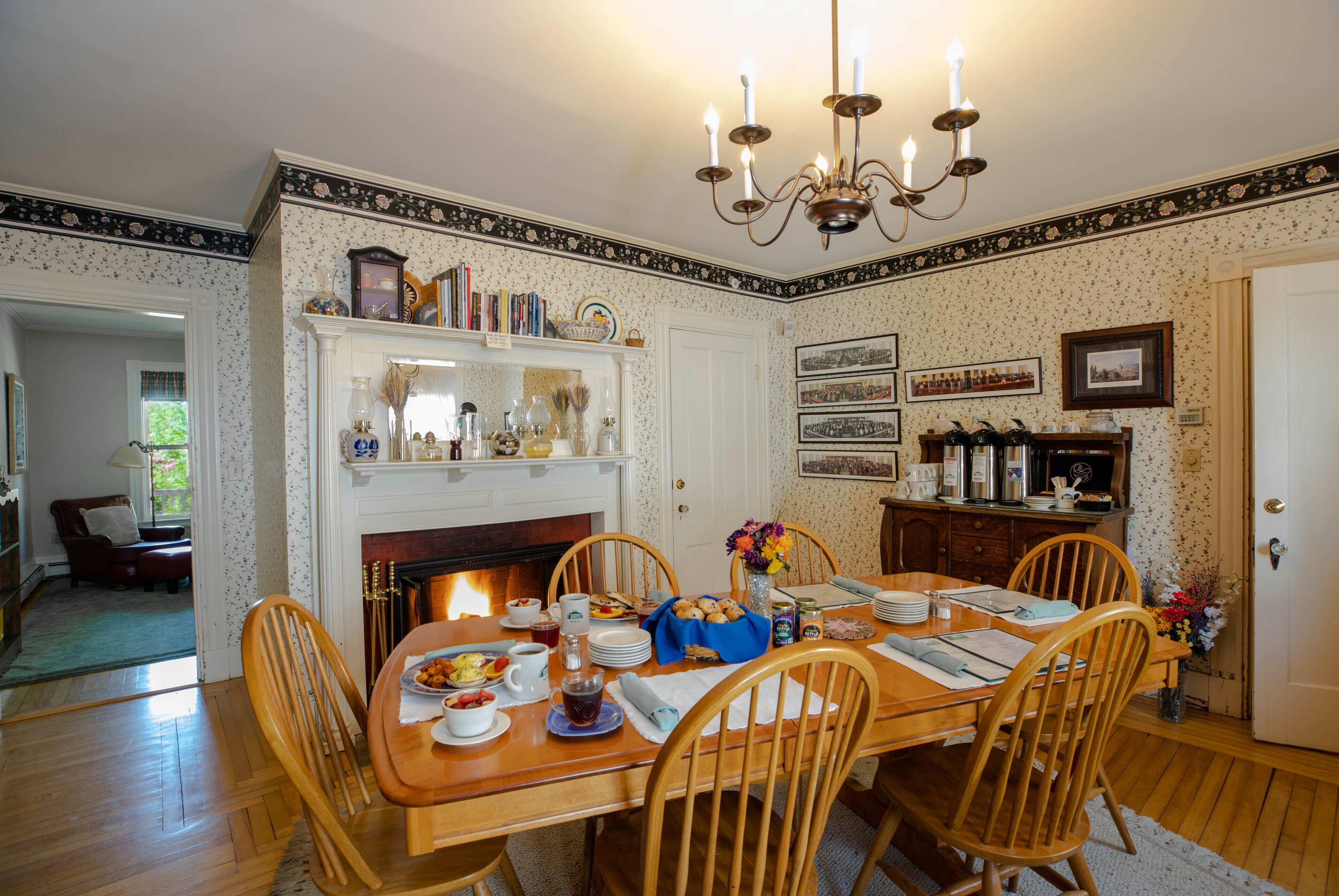 Un comedor con una mesa de madera dispuesta para cuatro personas, rodeada de sillas. Se observa una chimenea al fondo, y las paredes tienen un papel tapiz con diseño, además de una araña.