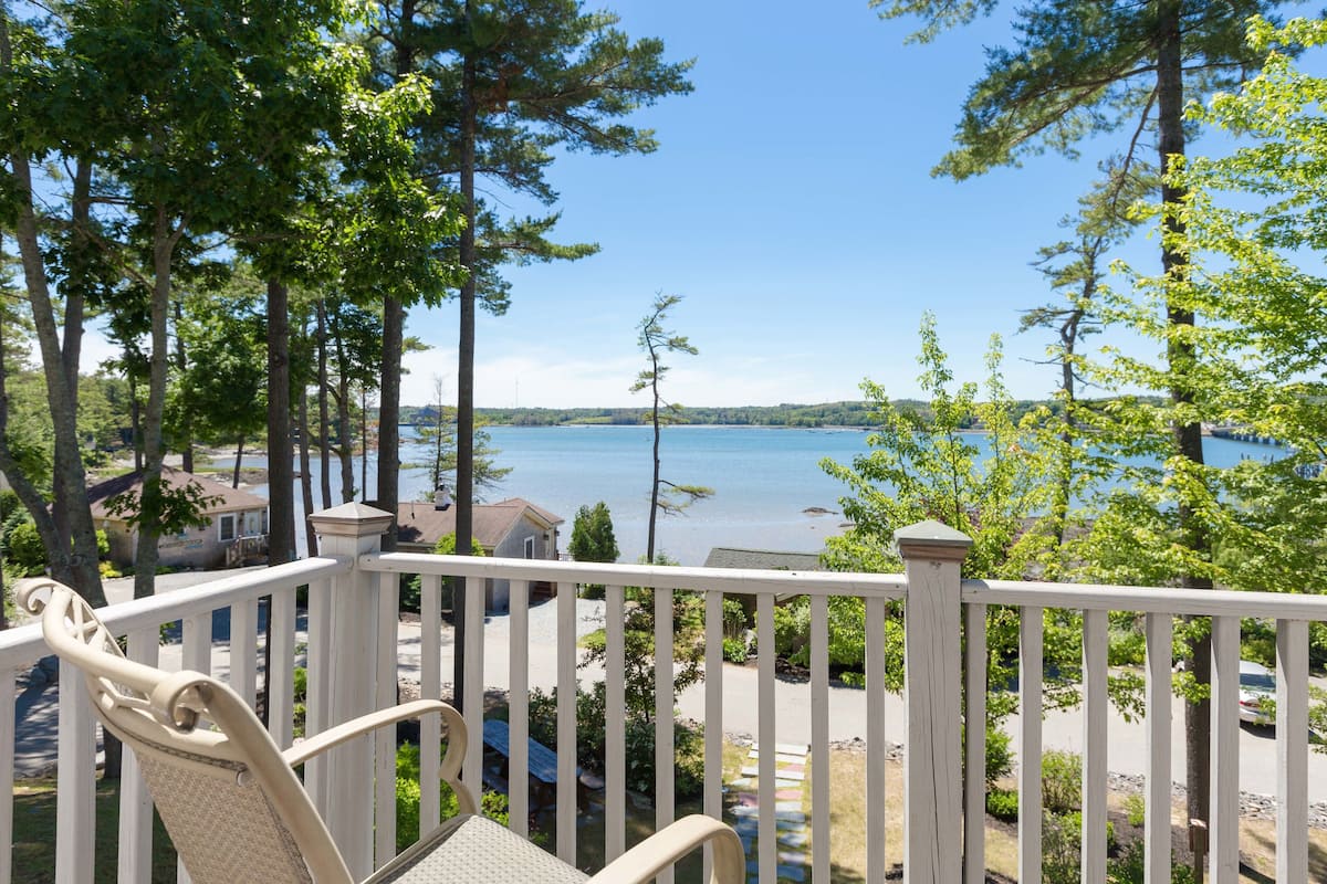 A wooden deck with a chair, overlooking a lake with boats and houses in the background.