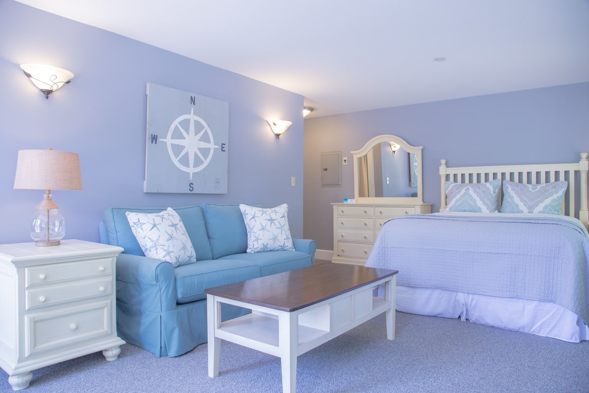 A bedroom with a blue sofa, a white dresser, a wooden nightstand, a bed with a white headboard, and a compass wall art piece.