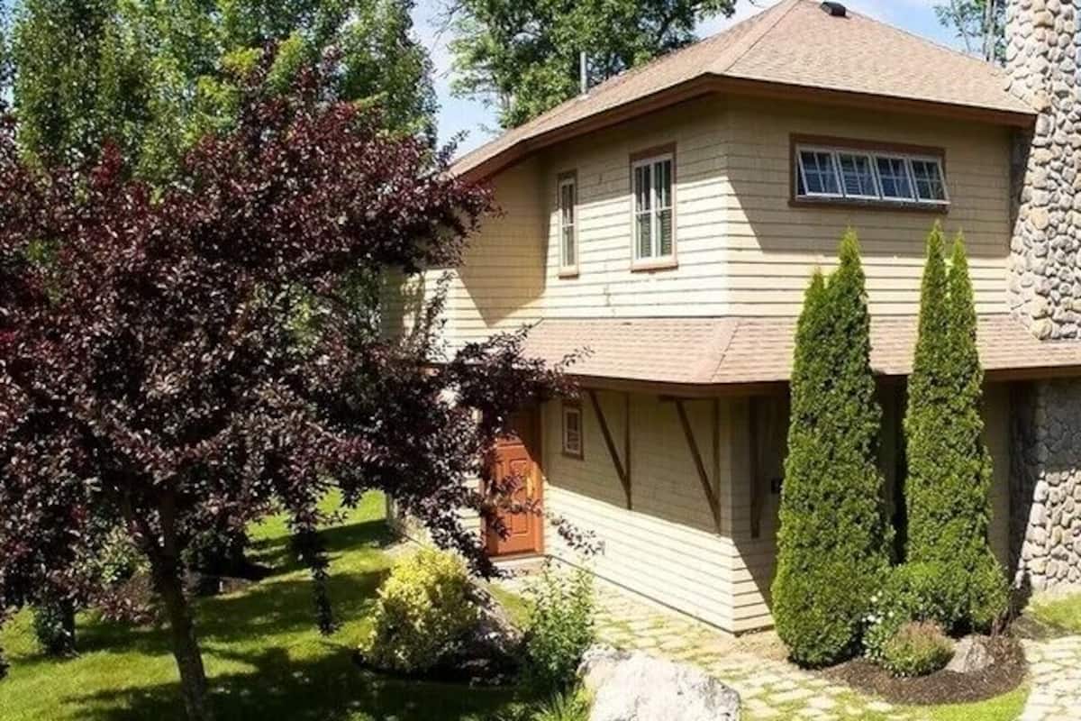 A two-story house with a stone chimney, surrounded by a well-maintained garden and trees.