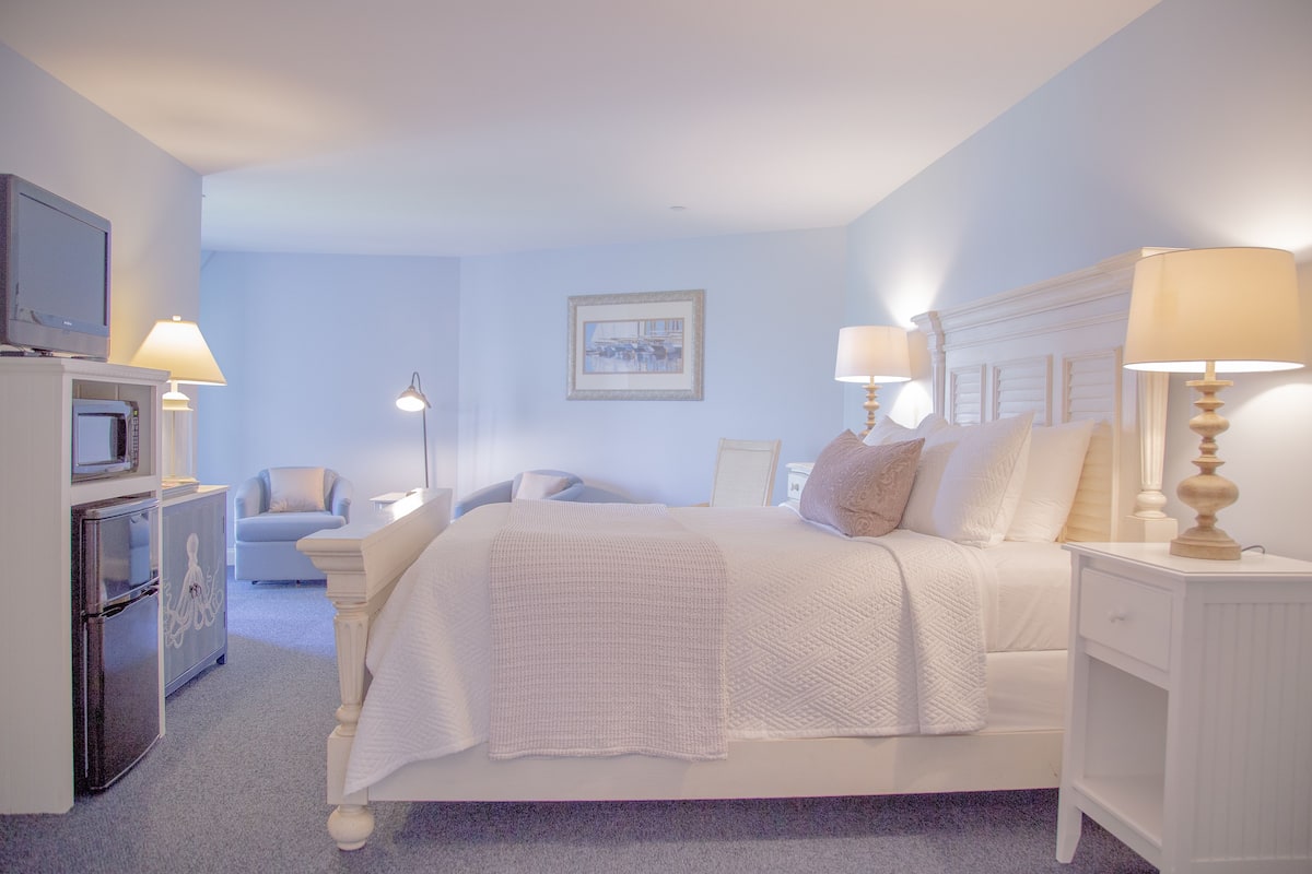 A hotel room with a bed, bedside tables, a TV, a chair, and a refrigerator.