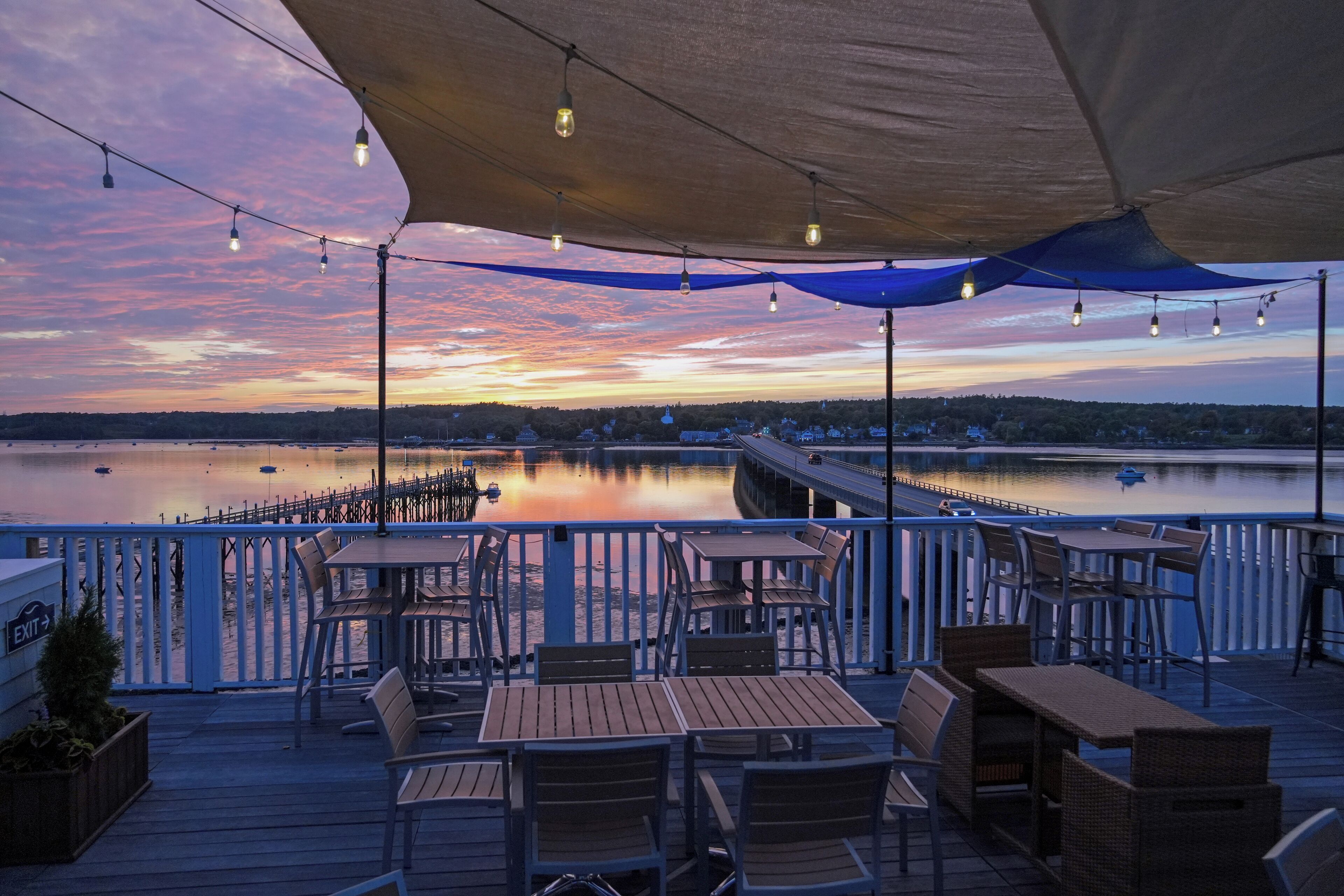 Een terras met tafels en stoelen met uitzicht op een rustig wateroppervlak bij zonsondergang.