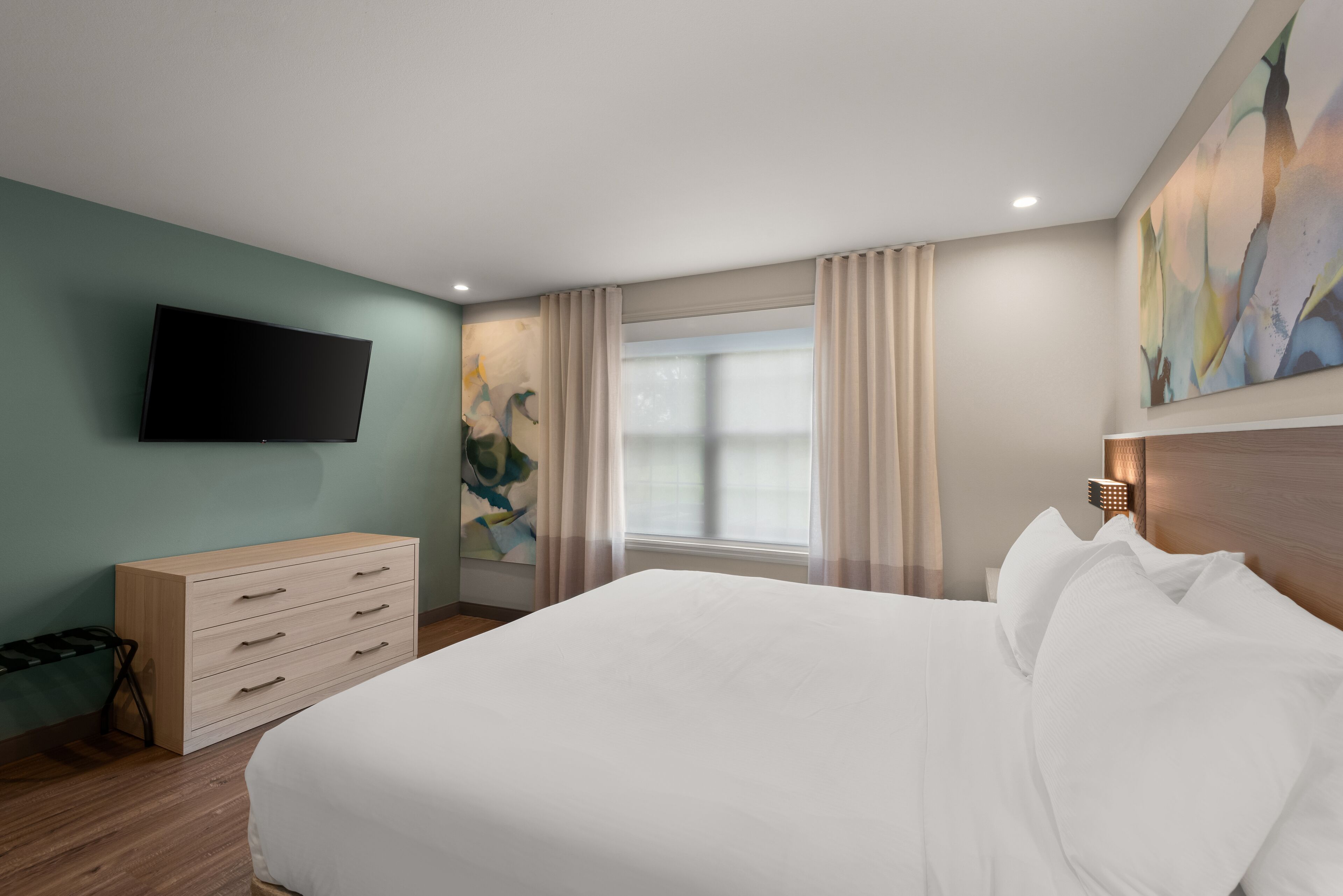 A hotel room with a bed, a flat-screen TV mounted on the wall, a wooden dresser, a window with curtains, and a painting on the wall.
