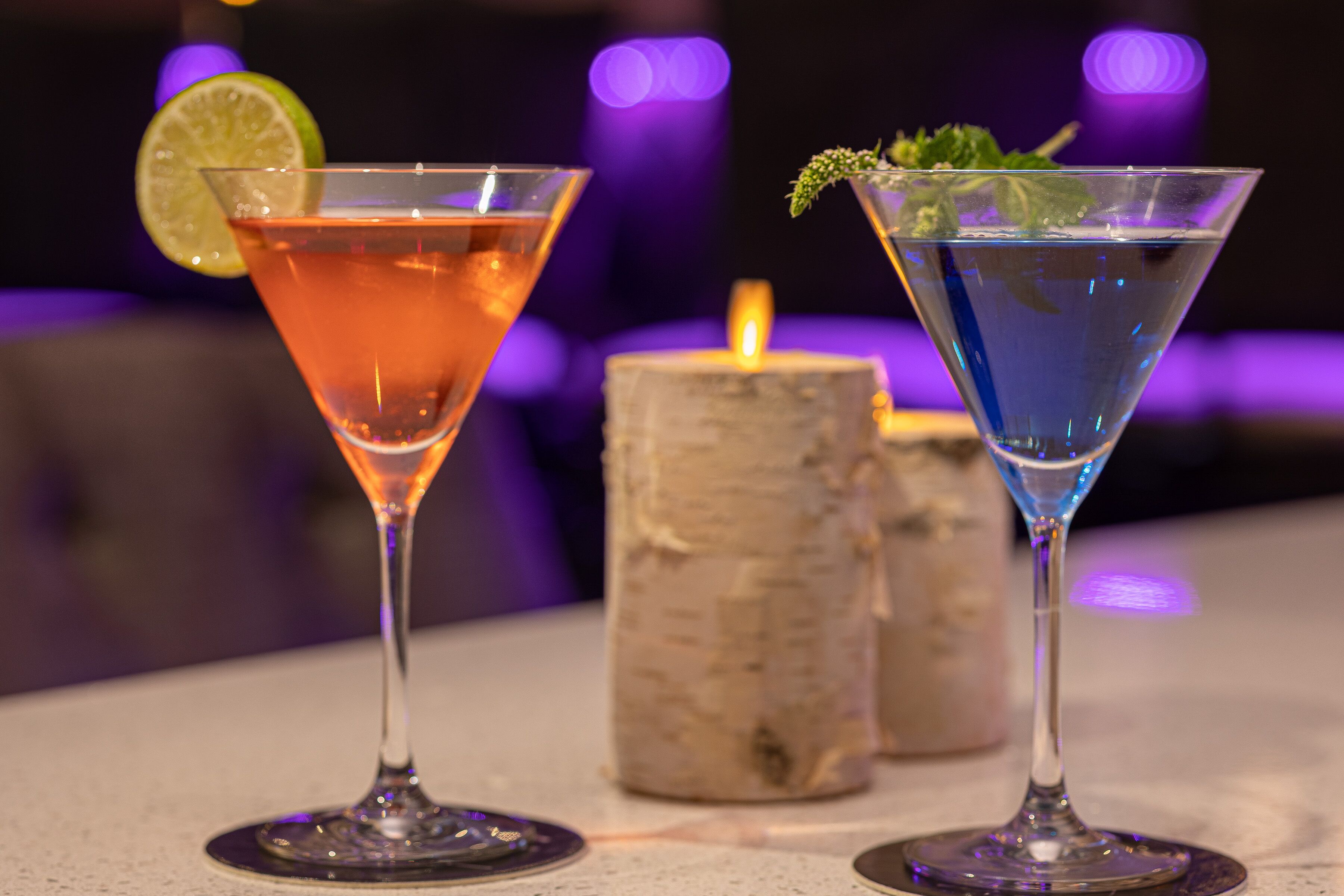 Two martini glasses with different colored drinks, a candle, and a blurred background with purple lighting.