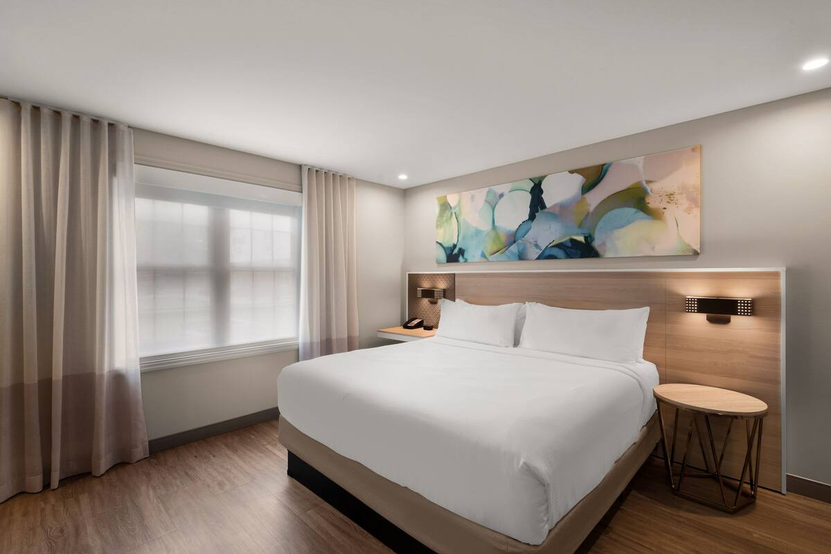 Una habitación de hotel moderna con una cama grande, una mesita de noche, una mesita redonda auxiliar, una lámpara de pared y un cuadro abstracto de gran tamaño.