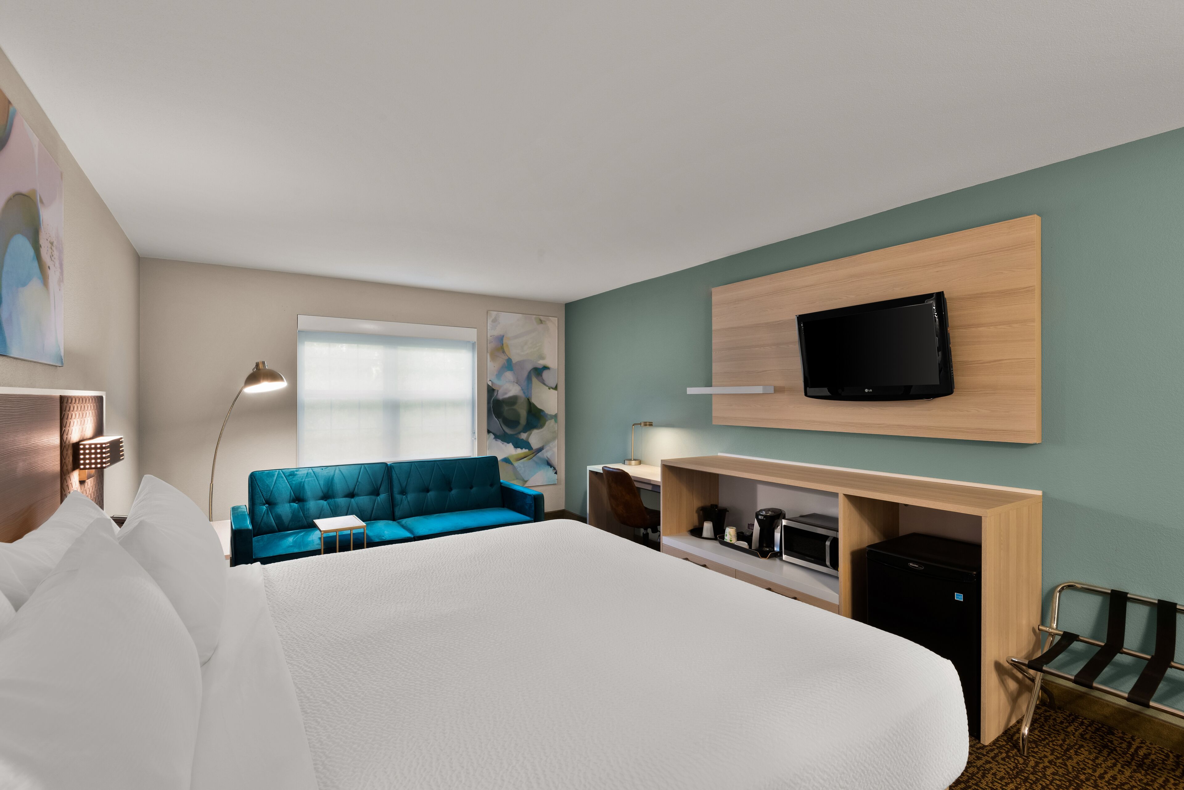 A modern hotel room with a large bed, a blue sofa, a desk with a computer, a flat-screen TV mounted on the wall, and a window with a view.