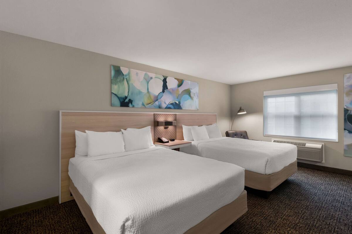 Habitación de hotel con dos camas, un cuadro abstracto grande en la pared, una ventana con persianas y un calentador fijado a la pared.