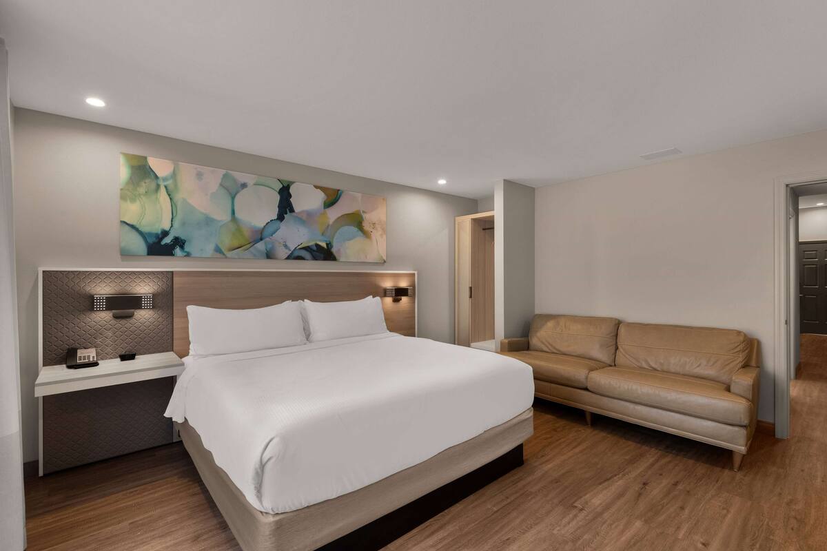 Un dormitorio moderno con una cama grande, una mesita de noche con lámpara, un sofá color beige y arte abstracto en la pared.