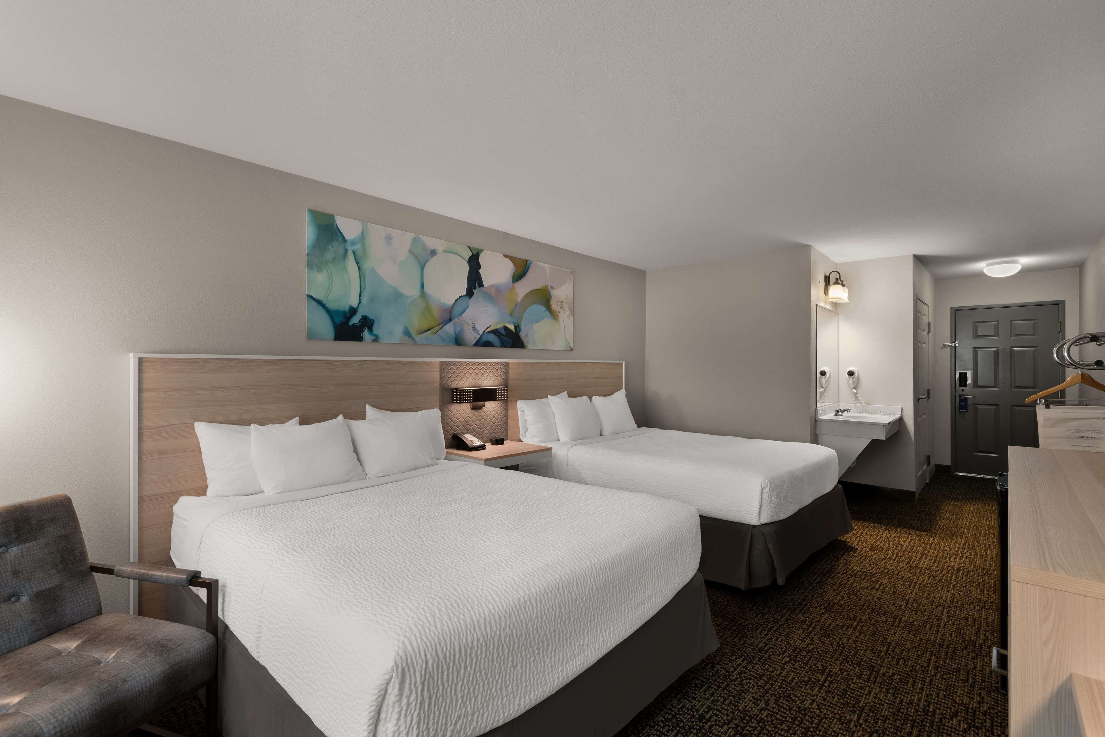 A hotel room with two beds, a desk, a chair, a painting on the wall, and a bathroom with a mirror and sink.