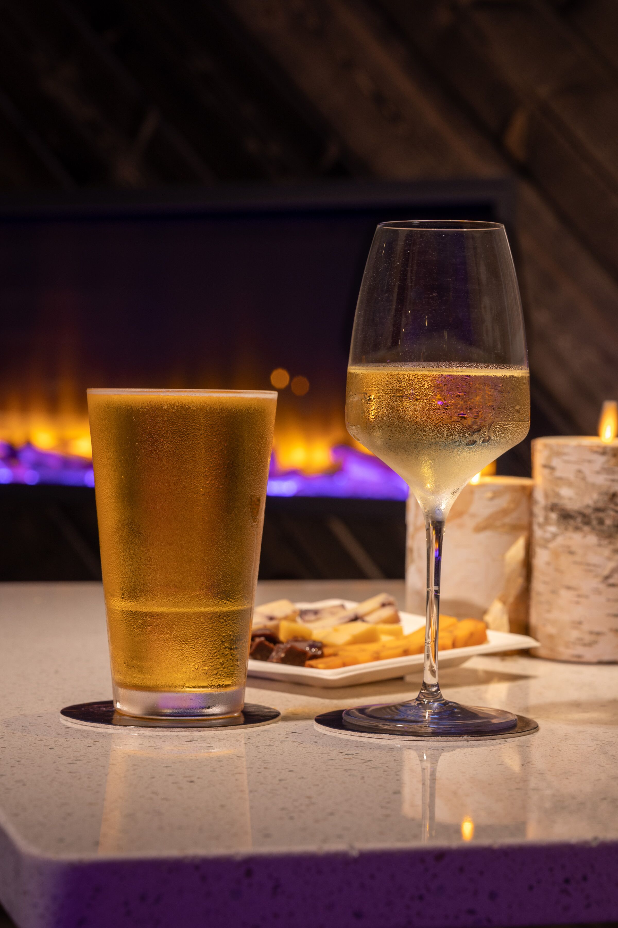 A glass of white wine and a tall glass of beer on a table with a candle and a plate of fries.