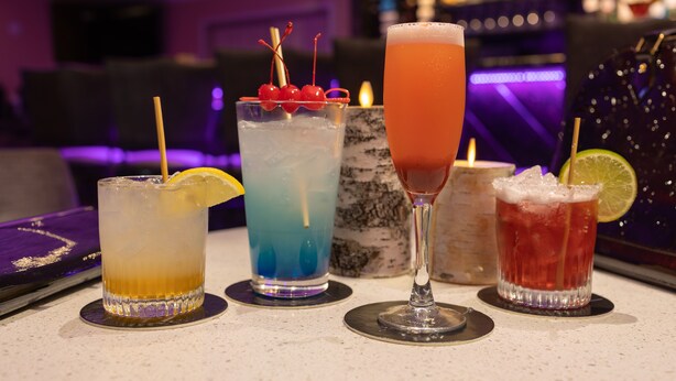 Quatre cocktails différents sont présentés sur le comptoir d’un bar, avec un arrière-plan flou représentant le bar et l’espace réservé aux clients.
