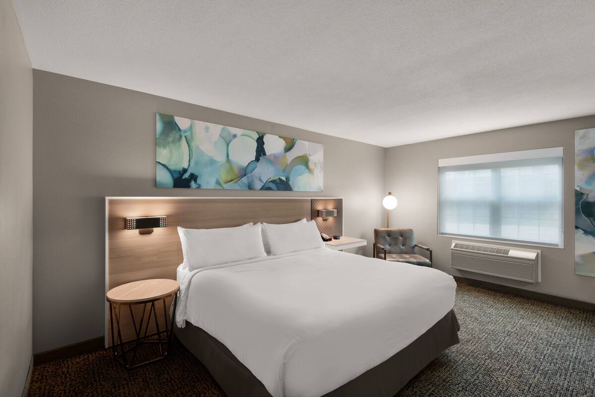 Habitación de hotel con una cama grande, una mesita redonda, una silla, una lámpara de pared y un amplio ventanal con persianas.