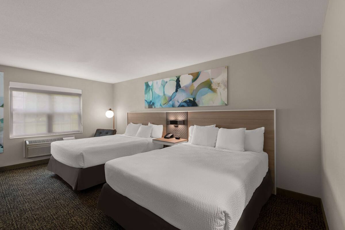 Habitación de hotel con dos camas, un cuadro abstracto grande, una mesita de noche con lámpara y una ventana con persianas.