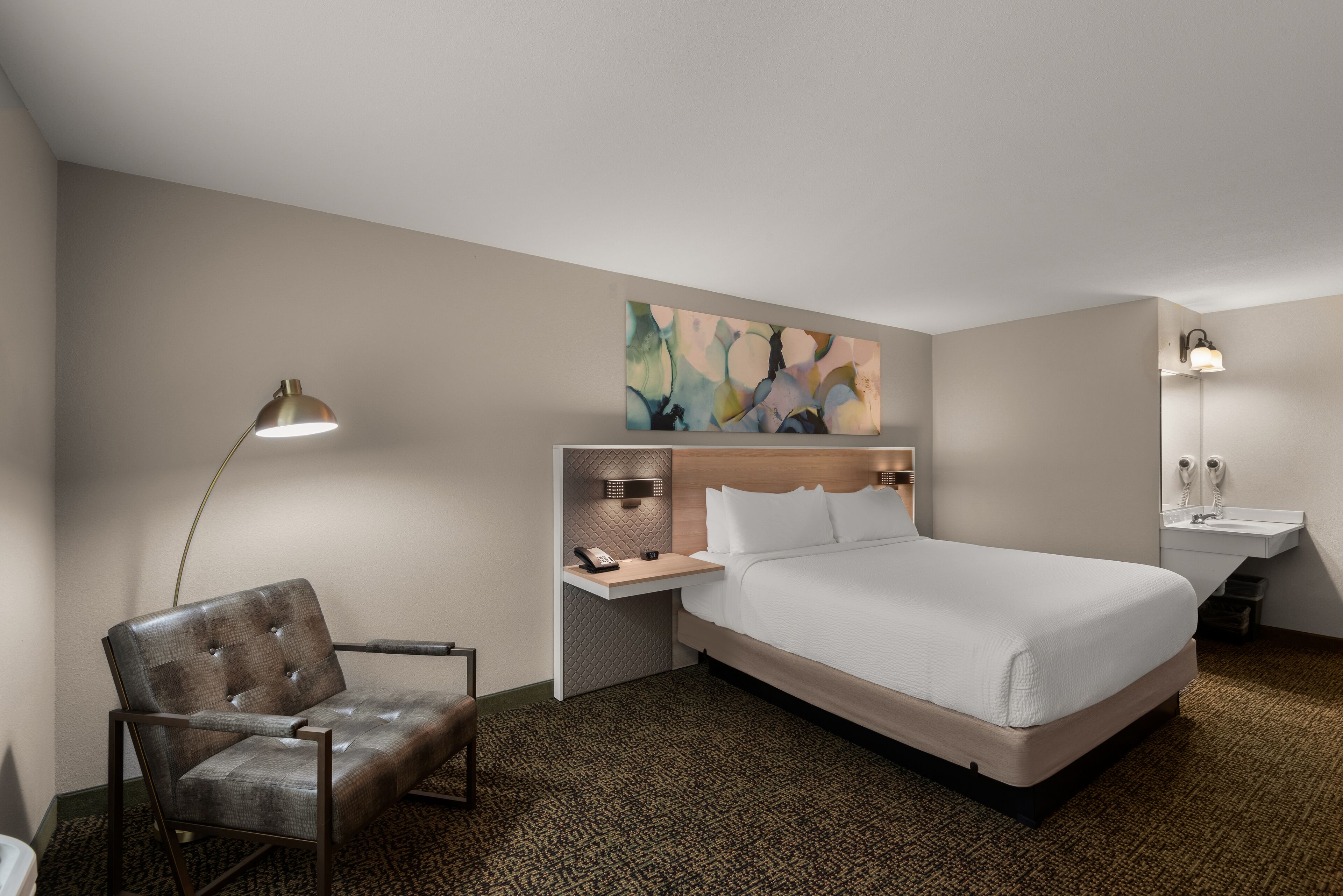Une chambre d’hôtel comprenant un lit, une chaise, un bureau avec une lampe, un tableau au mur et une salle de bain avec un lavabo et un miroir.