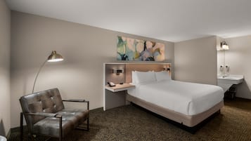 Une chambre d’hôtel comprenant un lit, une chaise, un bureau avec une lampe, un tableau au mur et une salle de bain avec un lavabo et un miroir.