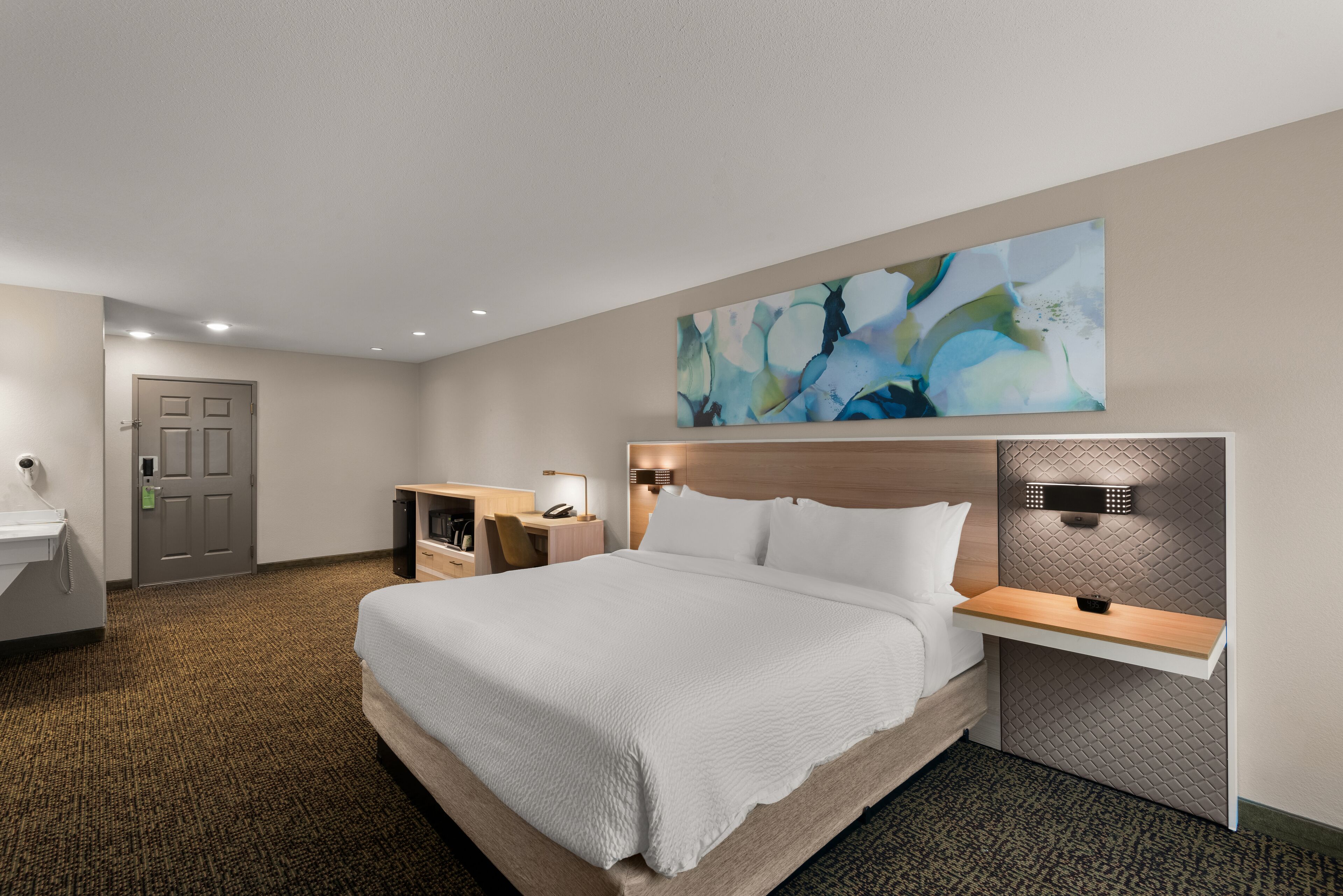 A hotel room with a large bed, a desk with a chair, a small table, and a wall-mounted shelf.