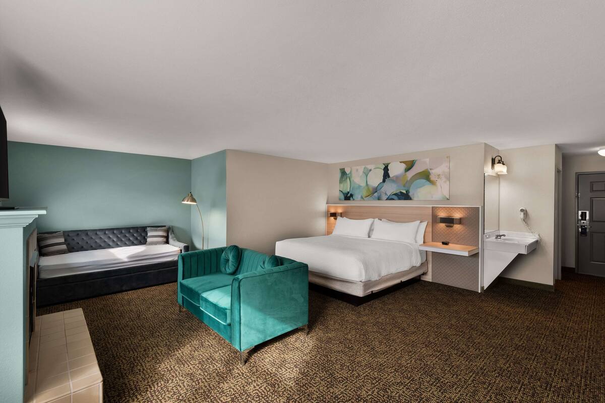 Una habitación de hotel con cama, sofá, sillón verde azulado, un cuadro en la pared, mesita de noche y lavamanos.