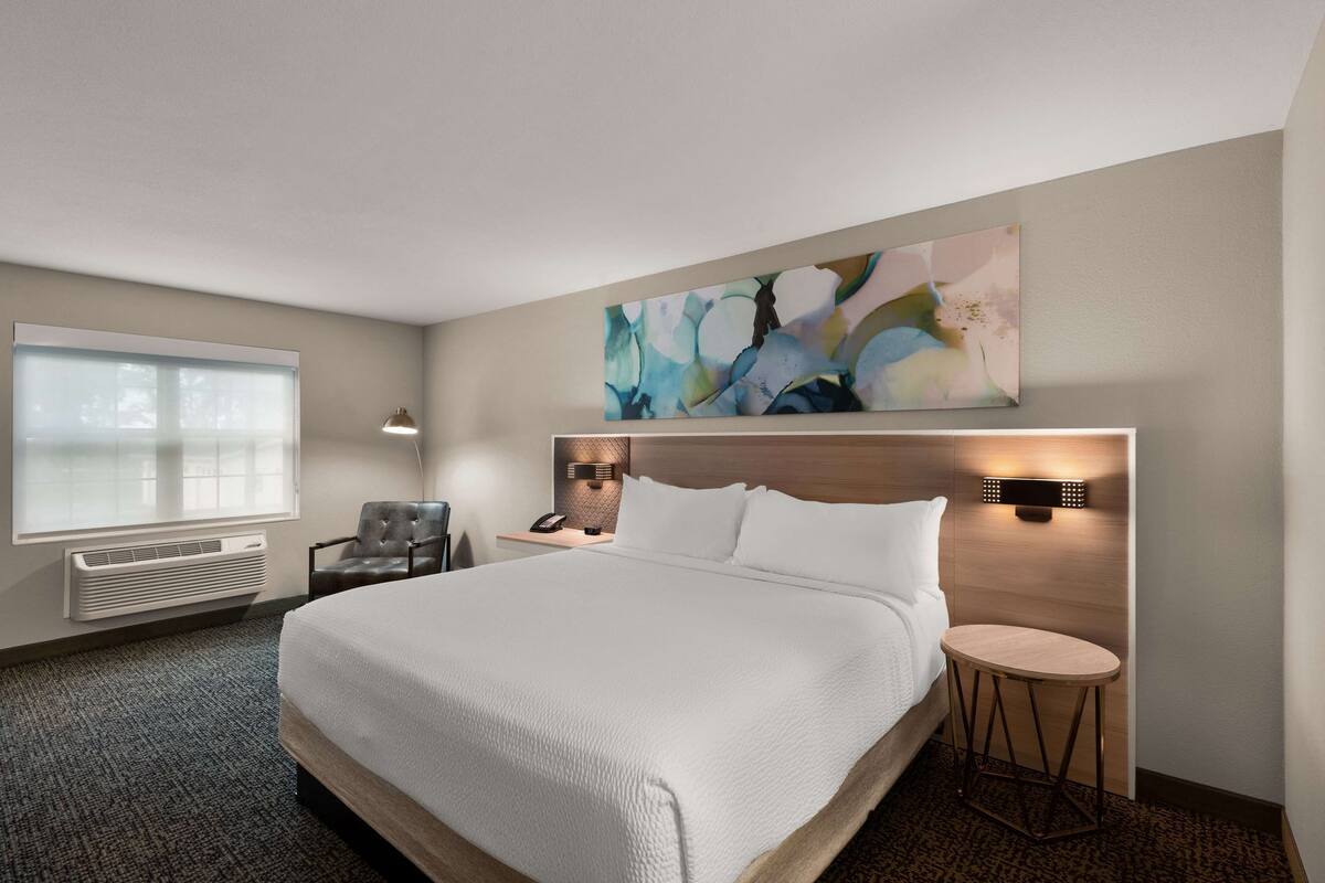 Una habitación de hotel con una cama grande, un escritorio con lámpara, una silla y un ventanal amplio.