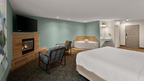 Habitación de hotel con cama, televisor montado en una chimenea de ladrillo, zona de estar con sillones, bañera y un baño pequeño con lavamanos y espejo.