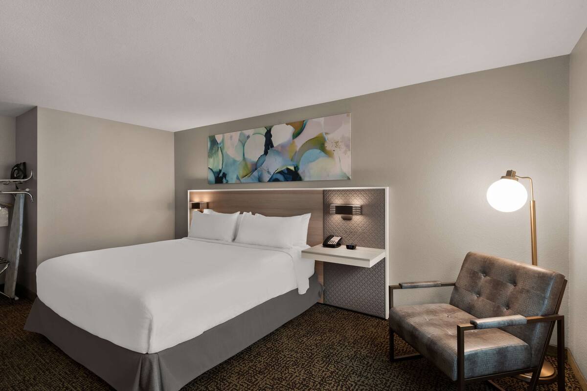 Una habitación de hotel con una cama grande, una mesita de noche con lámpara, una silla y un teléfono en la pared.