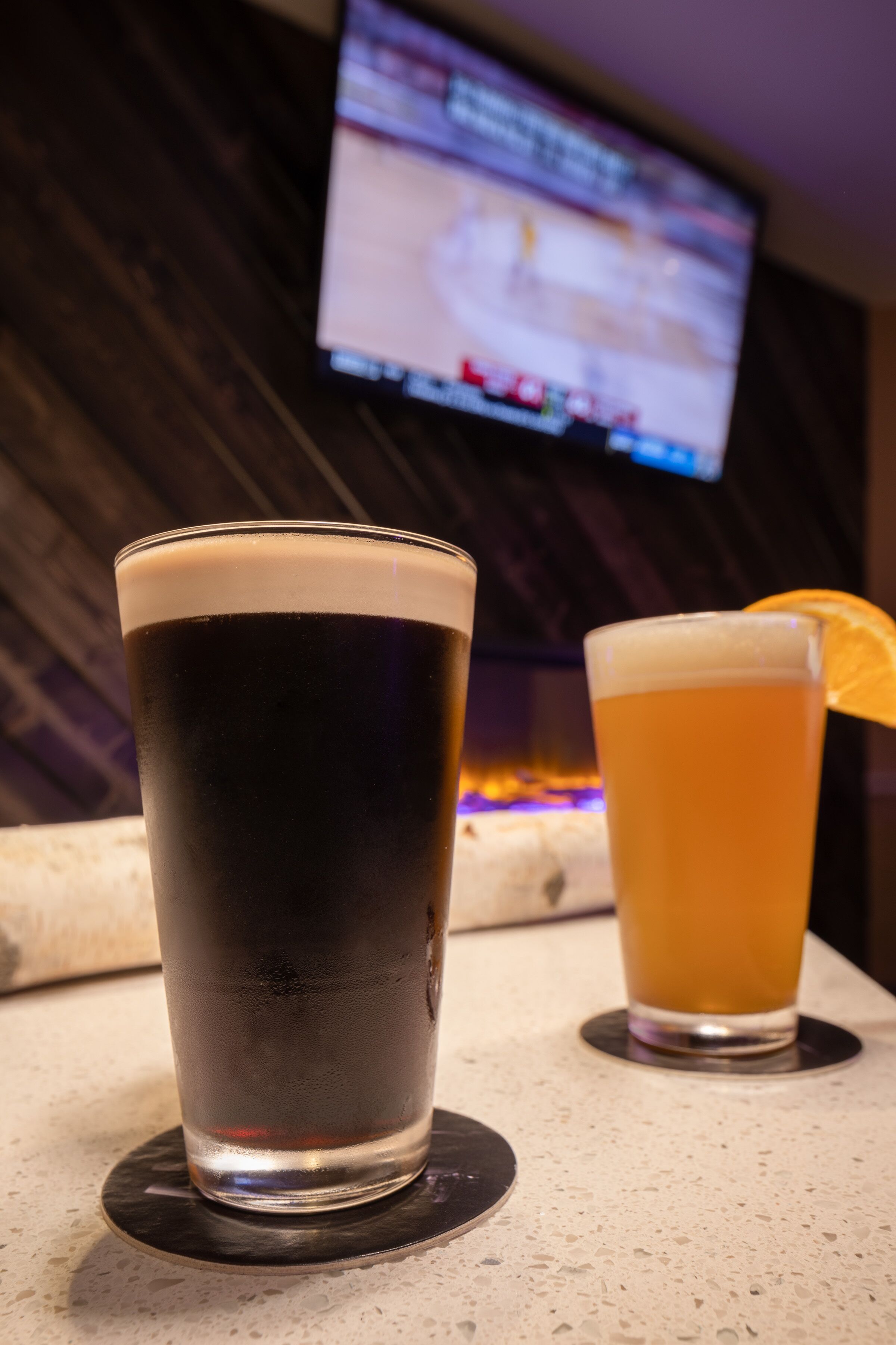 Deux verres de bière posés sur un dessous de verre, avec un téléviseur en arrière-plan.