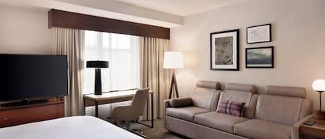 Habitación de hotel con cama, sofá, escritorio con lámpara, televisión y cuadros en la pared.