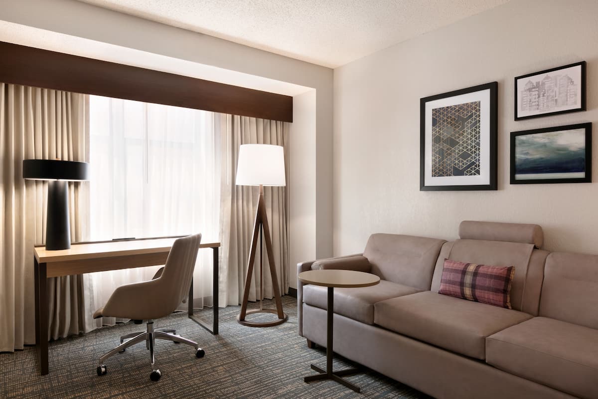 A hotel room with a desk, chair, sofa, lamp, and framed pictures on the wall.