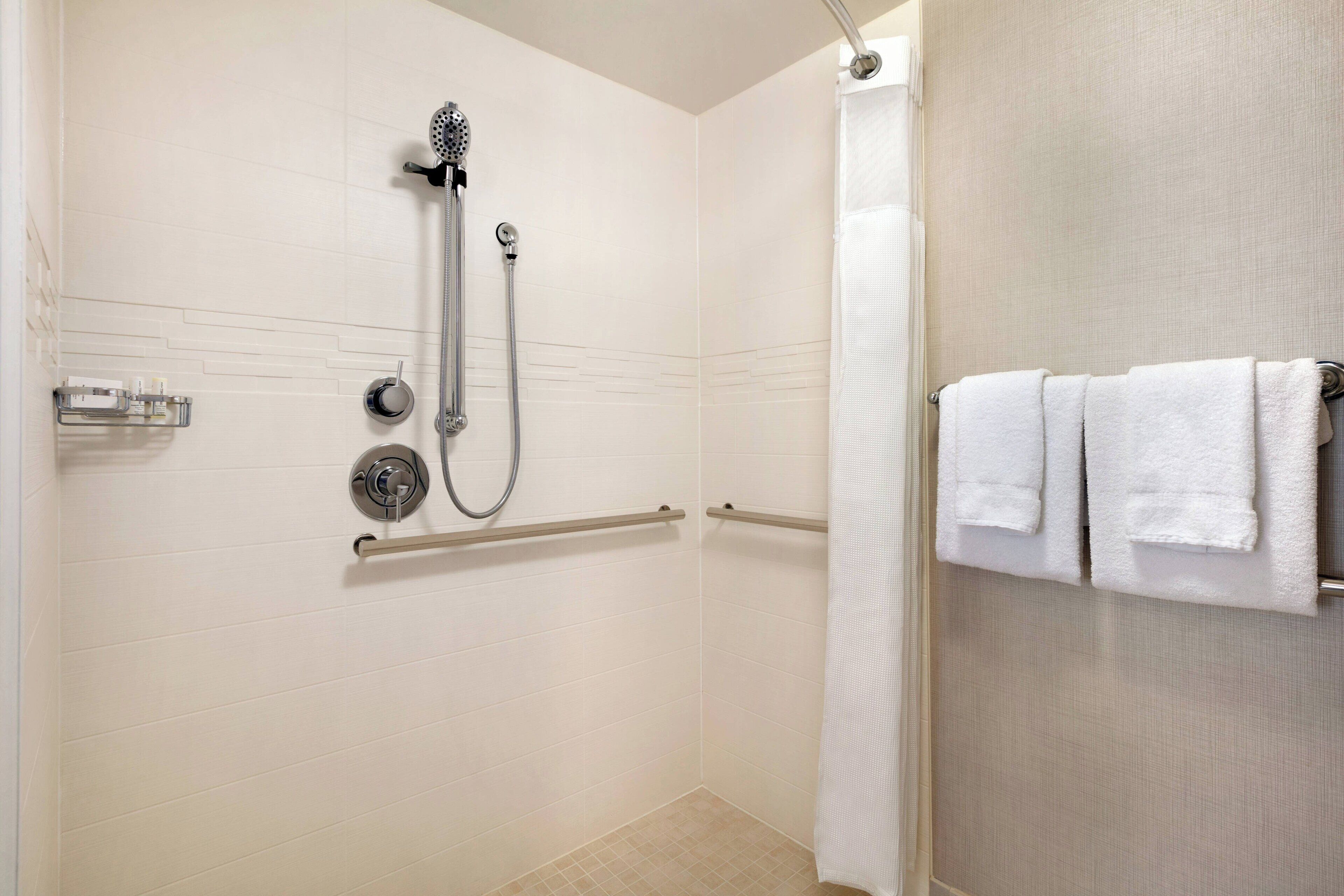 A shower with a handheld showerhead and a handheld hose, a towel rack with towels, and a handrail.