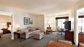 Habitación de hotel con sofá, sillón, televisión, escritorio y mesa de comedor.