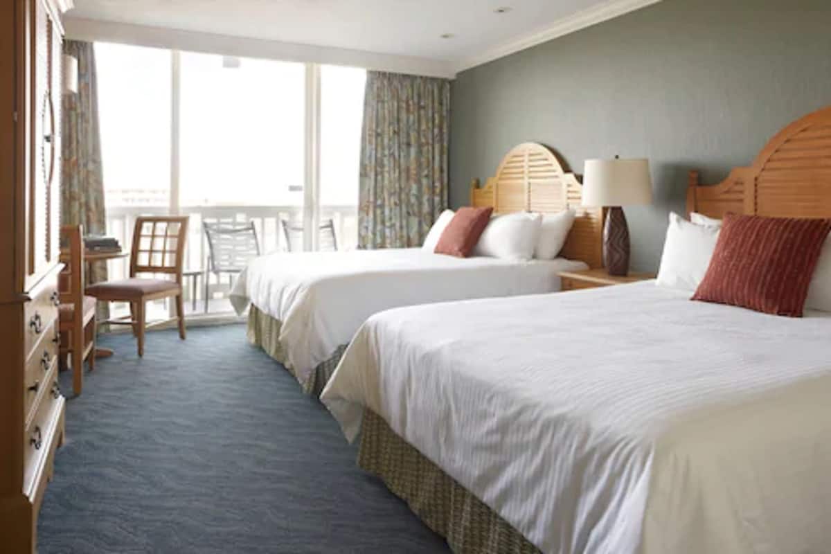 Una habitación de hotel con dos camas, un escritorio, una silla y un balcón con cortinas.