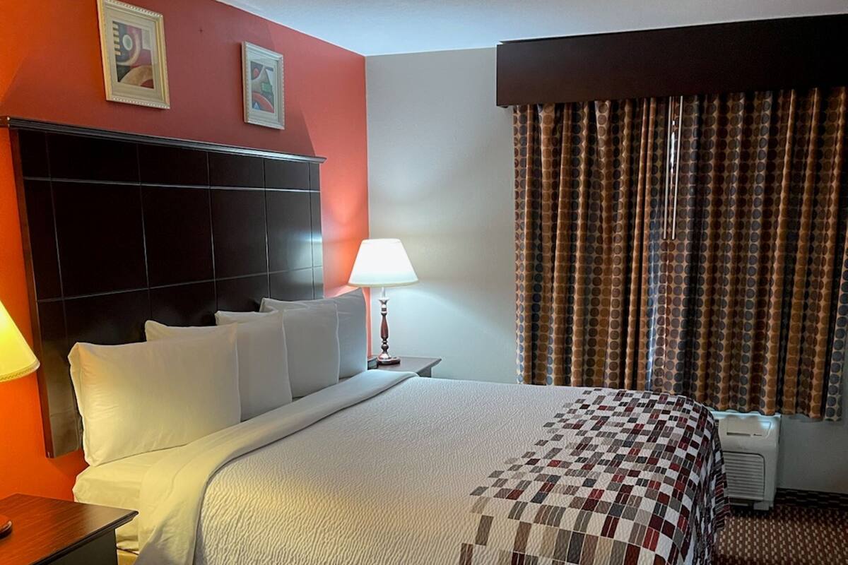 Ein Hotelzimmer mit einem großen Bett, einem Nachttisch, einer Lampe, einem an der Wand montierten Fernseher und einem Fenster mit Vorhängen.