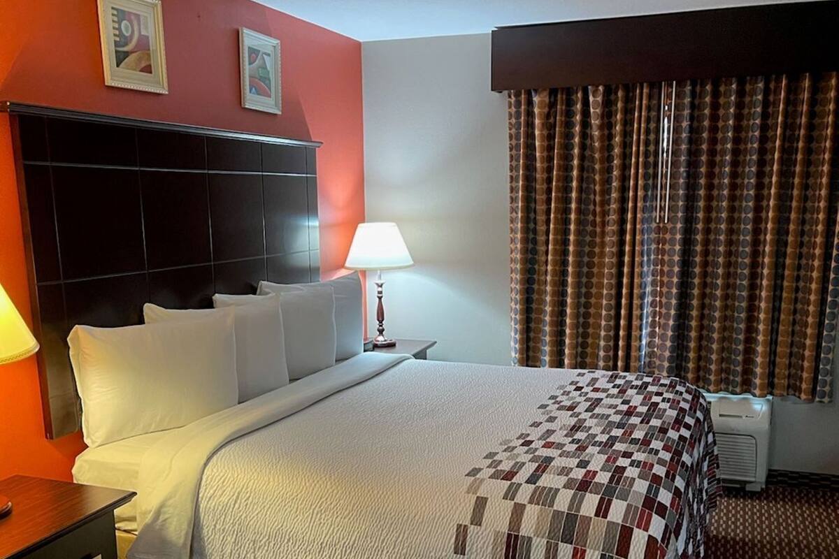 Ein Hotelzimmer mit Bett, Nachttisch, Lampe, Fenster mit Vorhängen und Kopfteil.