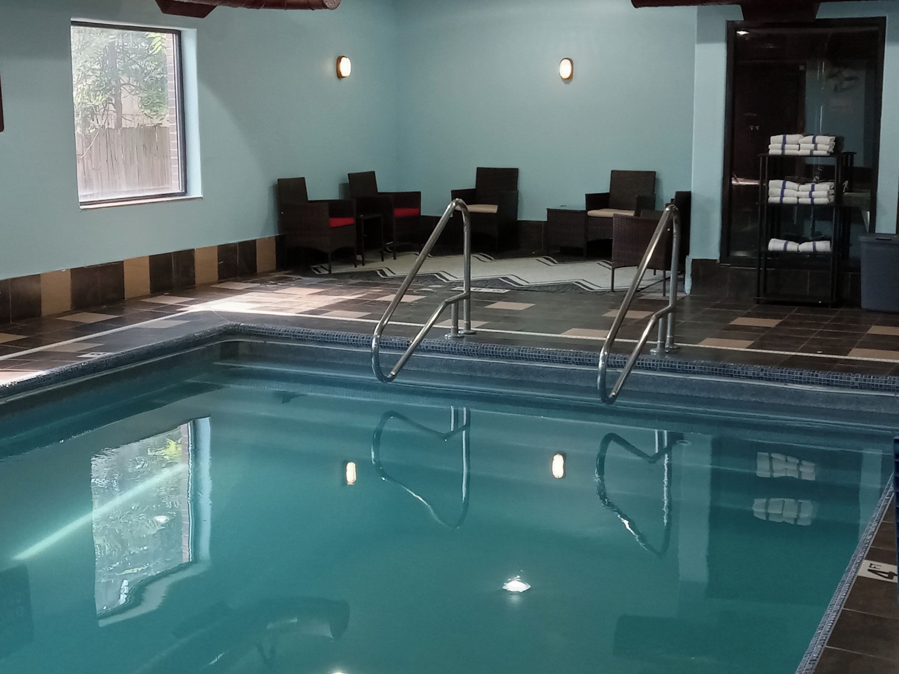 An indoor swimming pool with a tiled floor, a ladder, and seating area with chairs.