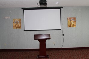 A conference room with a projector screen, a podium, and two paintings on the walls. - Red Roof Inn & Suites Detroit-Melvindale/ Dearborn (Melvindale)