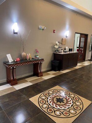 A reception area with a checkered floor, a decorative vase, a small table with a sign, and a coffee station. - Red Roof Inn & Suites Detroit-Melvindale/ Dearborn (Melvindale)