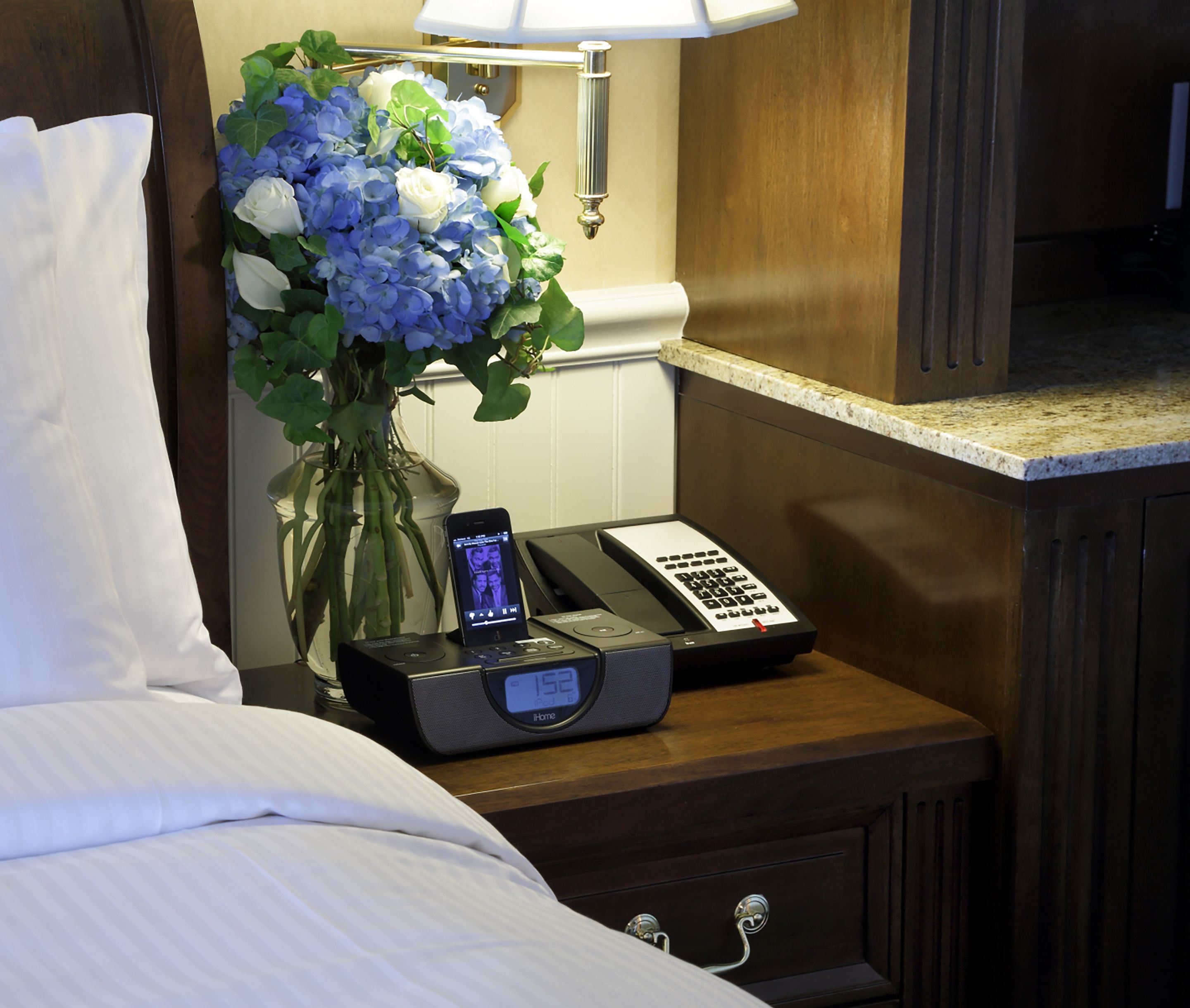 A hotel room with a bed, a bedside table with a lamp, a vase with flowers, a phone, and a digital clock.