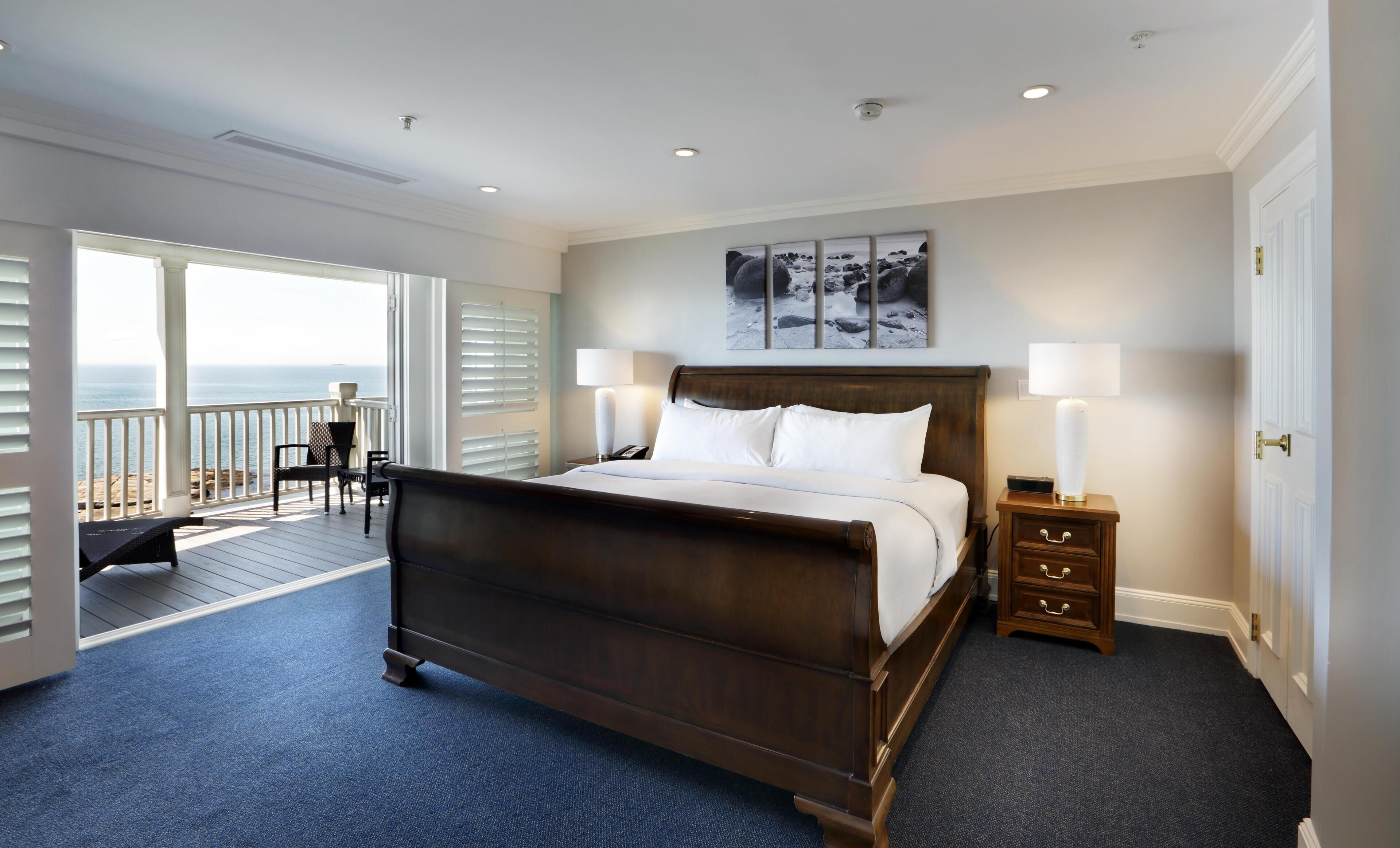 A hotel room with a large bed, two bedside tables, a view of the ocean, and a balcony with chairs.