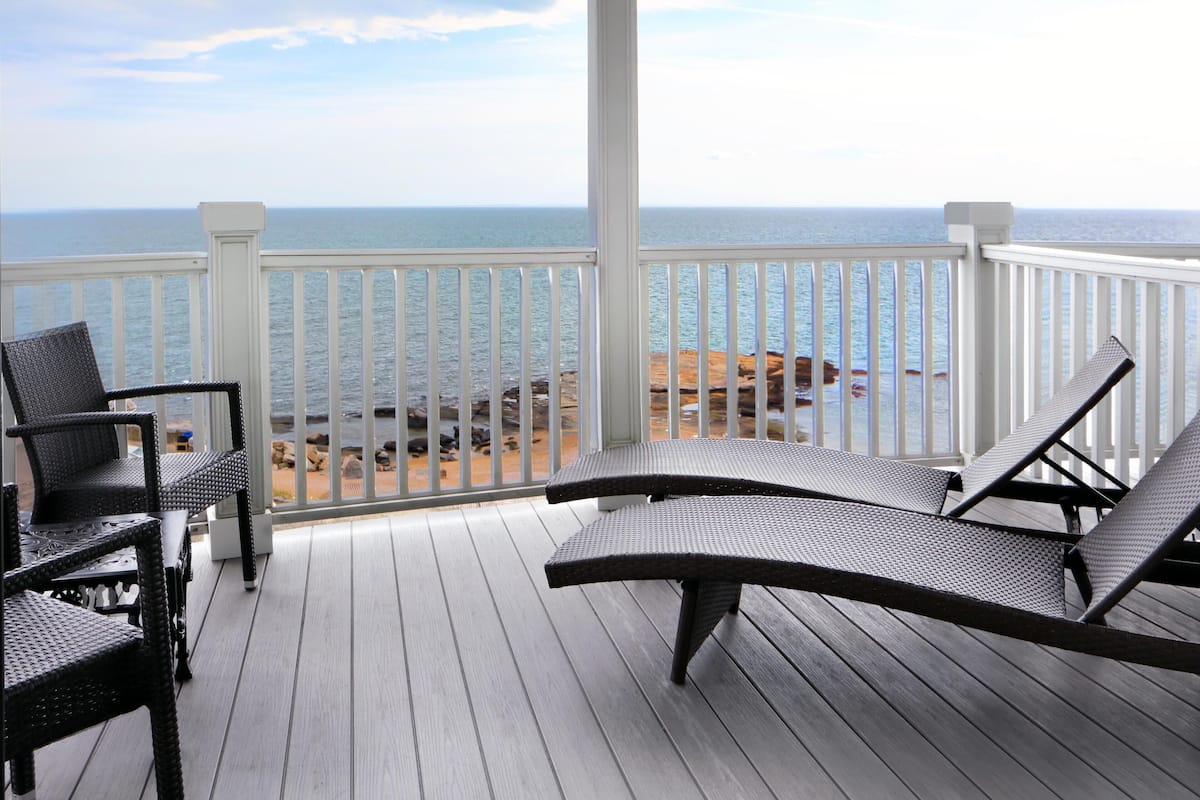 A covered deck with two lounge chairs and a table, overlooking the ocean.