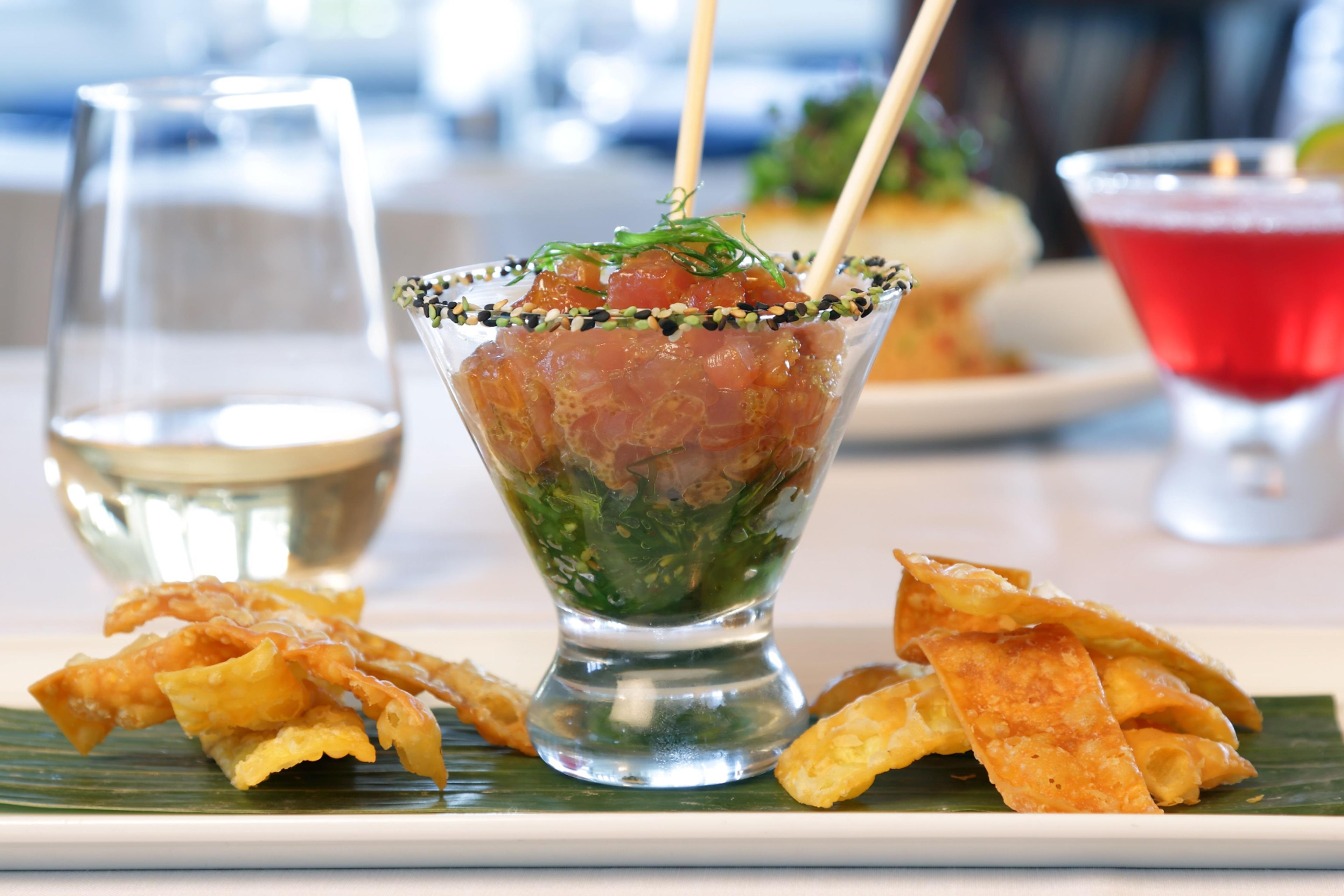 Un vaso con un plato de mariscos adornado con hierbas verdes y semillas de sésamo, acompañado de bocadillos fritos sobre una hoja de plátano y una bebida roja al fondo.