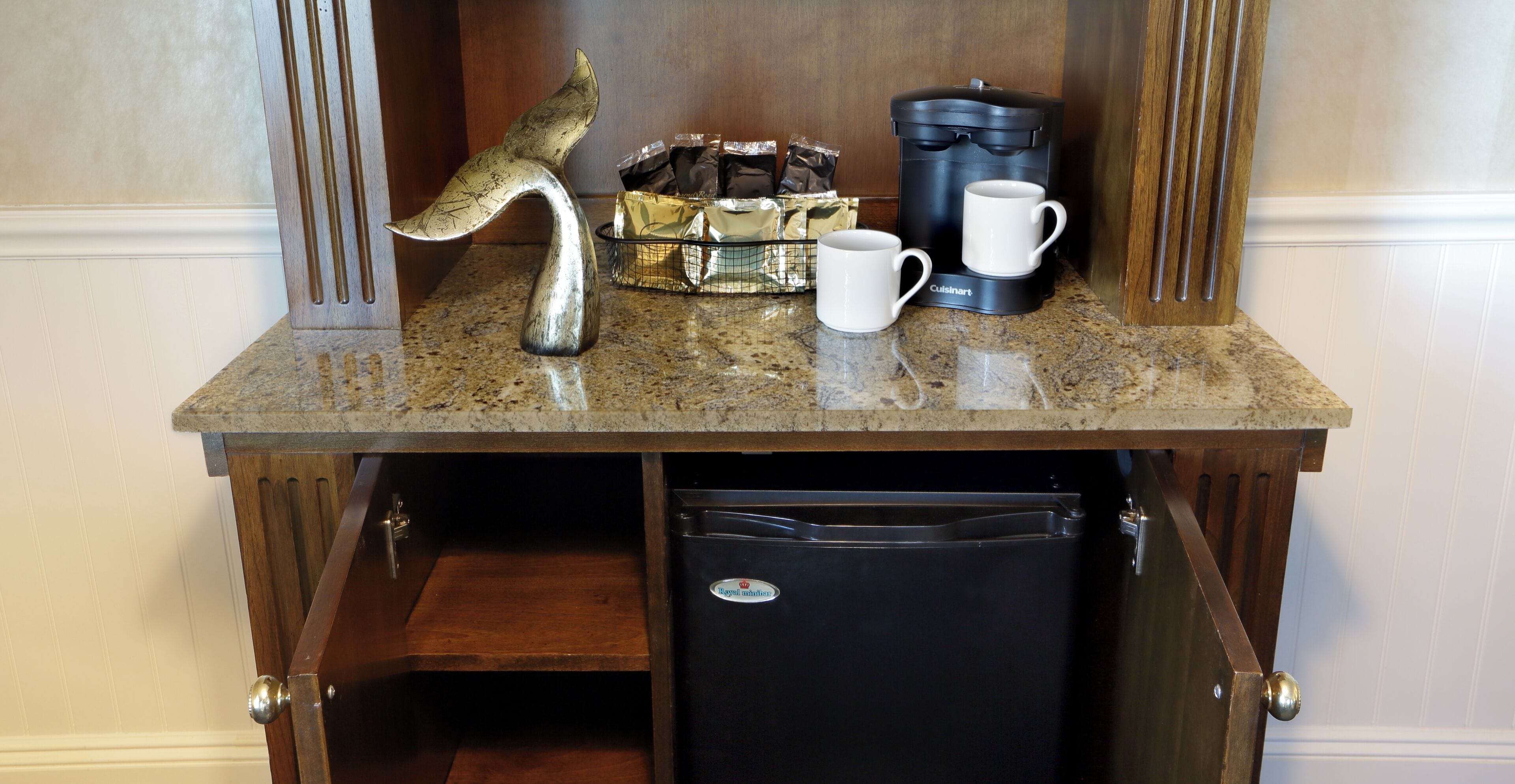 Una encimera de cocina con una cafetera, dos tazas, una escultura metálica decorativa y un refrigerador.