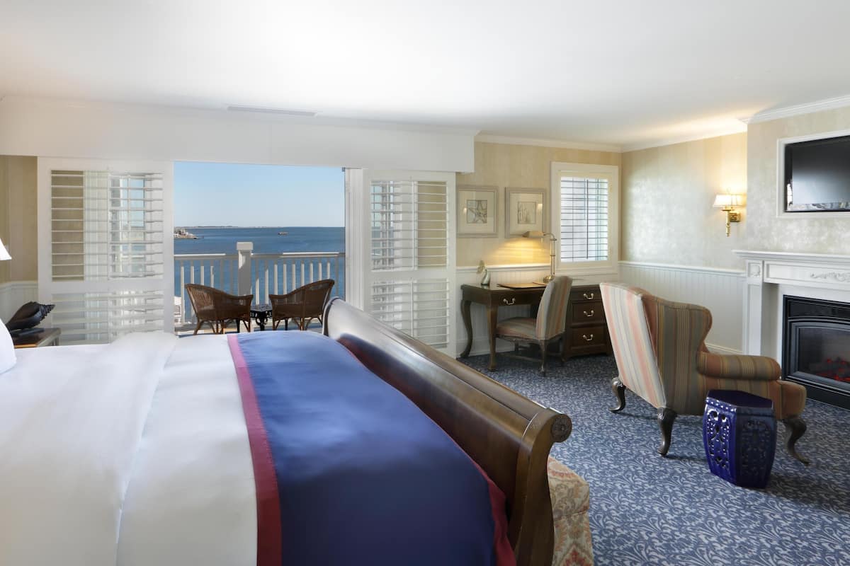A hotel room with a large bed, a desk, a chair, a fireplace, and a view of the sea.