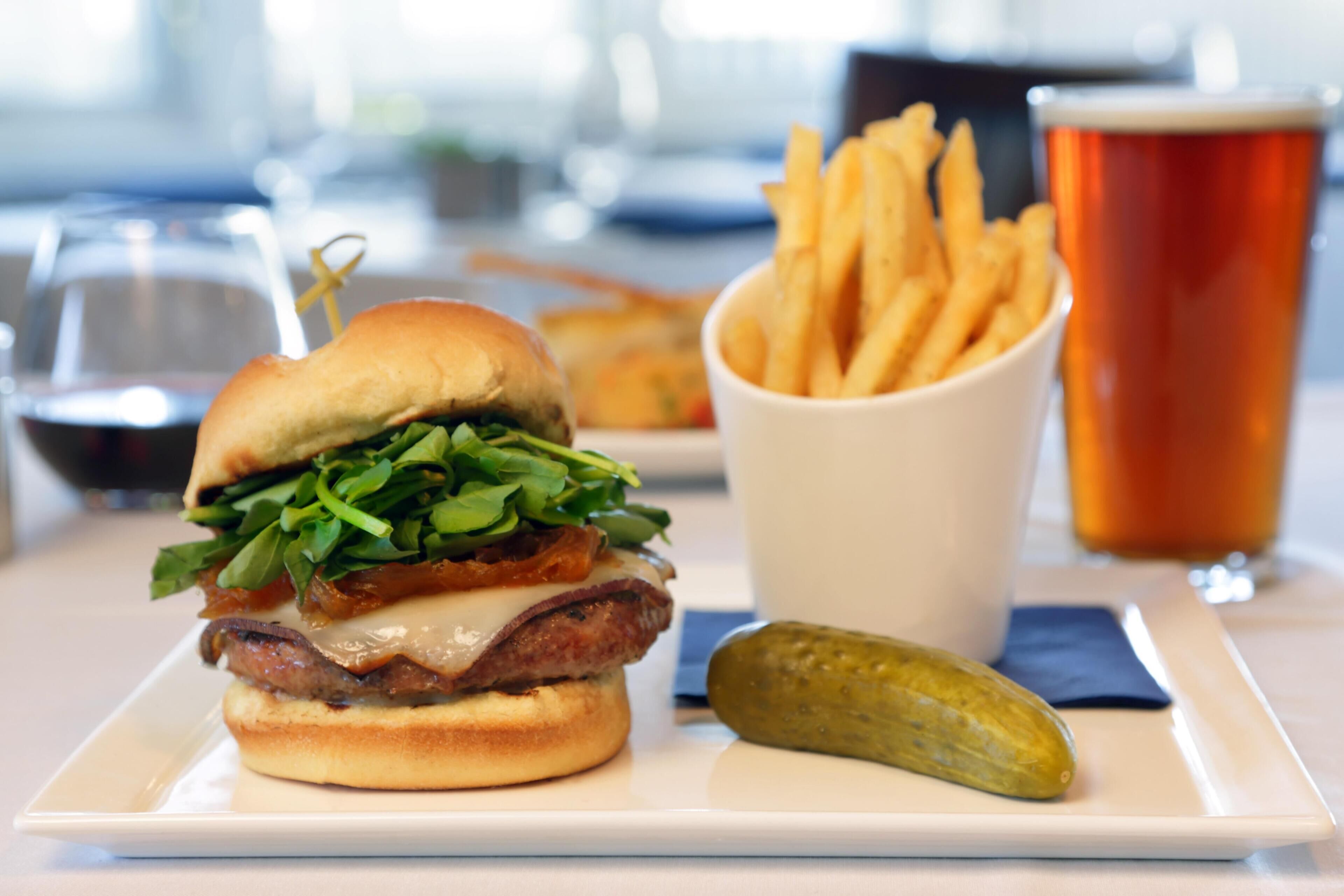 Un hamburger avec des frites et un cornichon, le tout servi sur une assiette blanche, accompagné d’un verre de bière et d’une tasse de café.