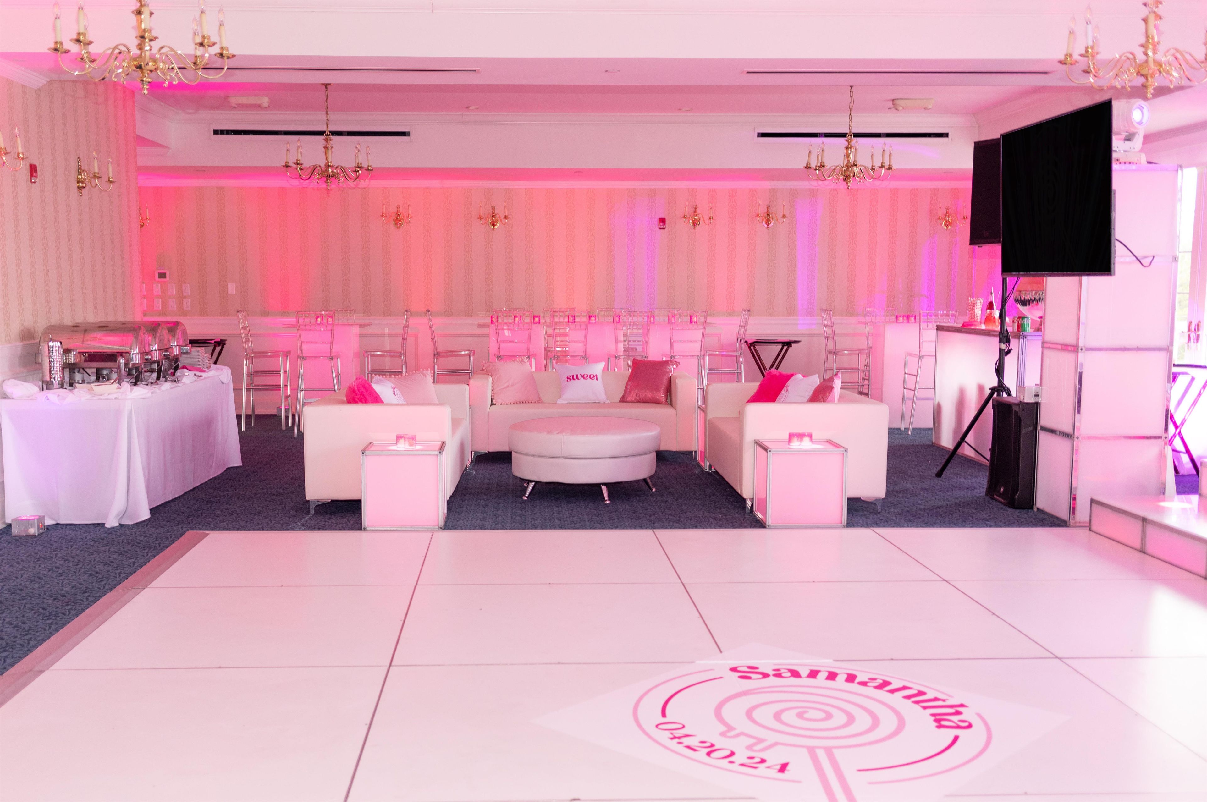 A room with a white floor, pink walls, and a chandelier. There are two white sofas, a small table, and a bar area with a TV.