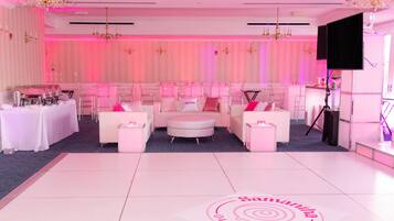 A room with a white floor, pink walls, and a chandelier. There are two white sofas, a small table, and a bar area with a TV.
